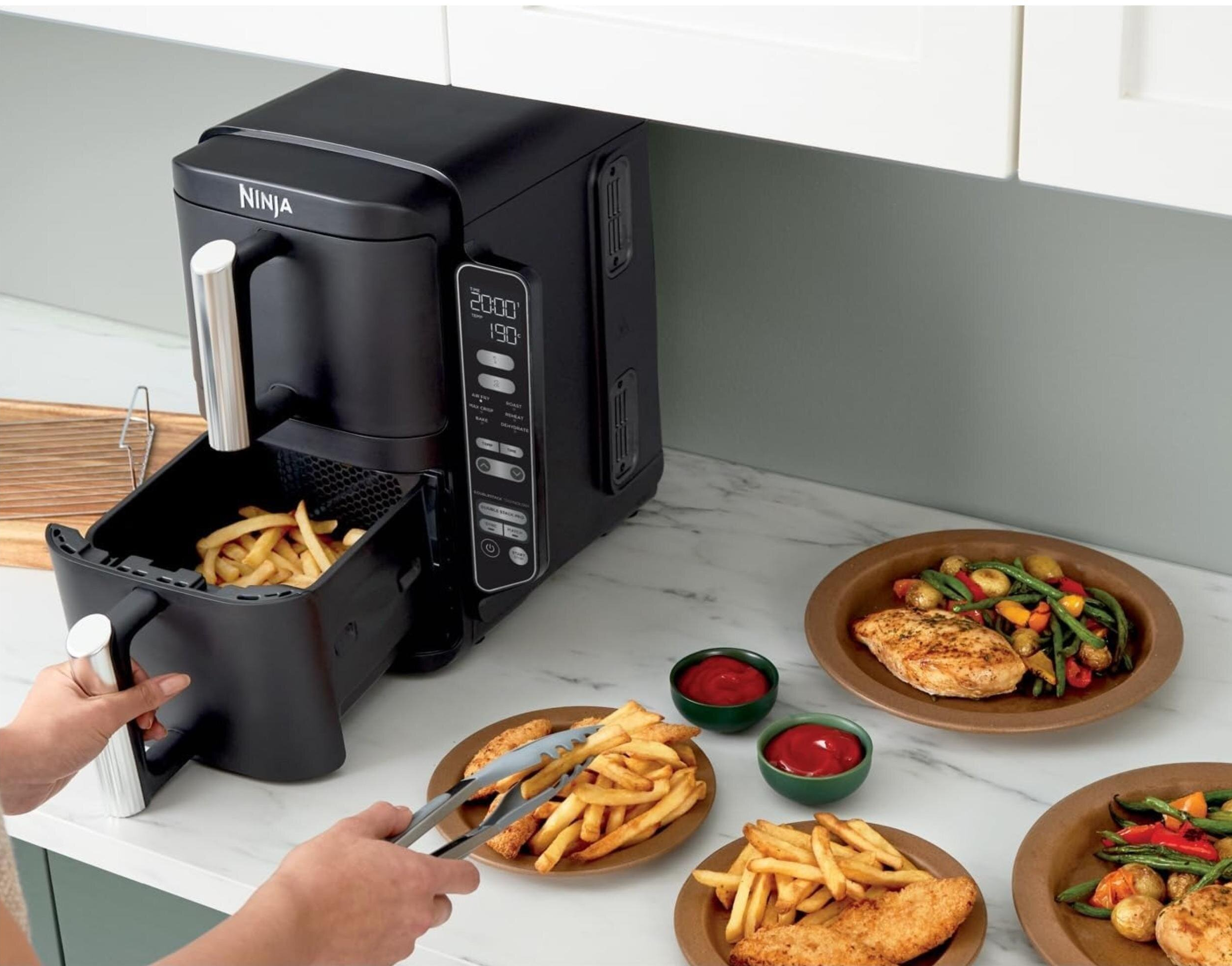 Czarna frytkownica powietrzna z frytkami, osoba trzymająca tacę i talerze z jedzeniem.