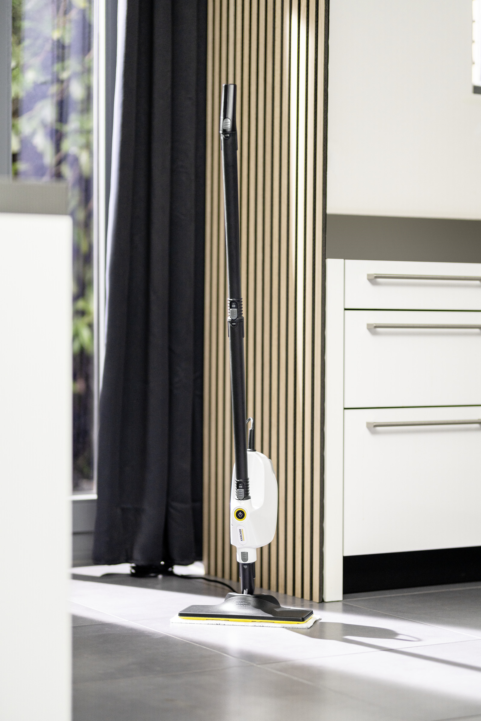 Czarny i biały mop parowy stoi pionowo w pokoju, w pobliżu okna i drewnianych paneli.