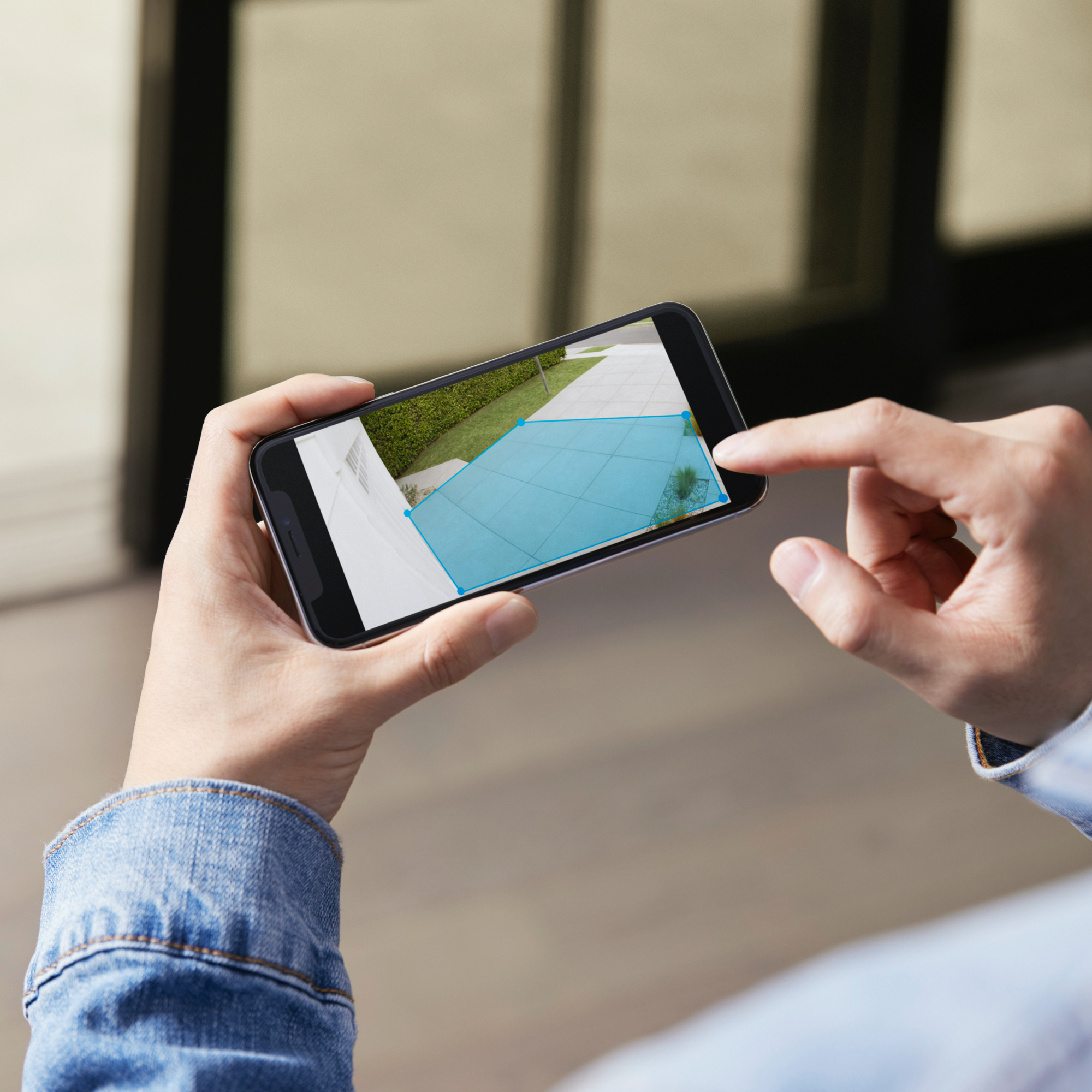 Hände einer Person, die ein Telefon halten. Der Bildschirm zeigt eine Ansicht einer Terrasse und einen blau markierten Bereich.