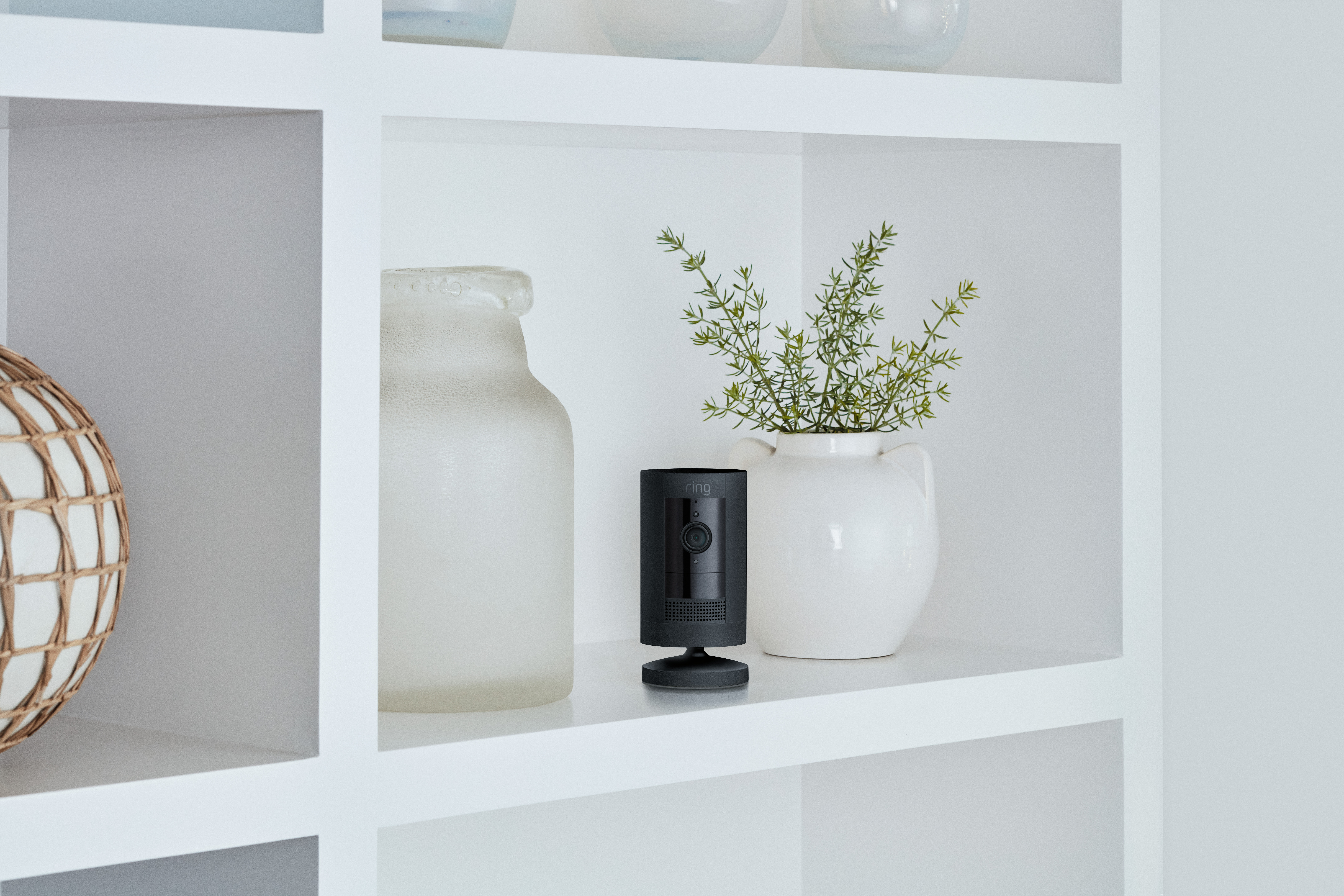 Weiße Regale mit schwarzer Überwachungskamera, Vase und Pflanze. Der Hintergrund ist eine hellgraue Wand.