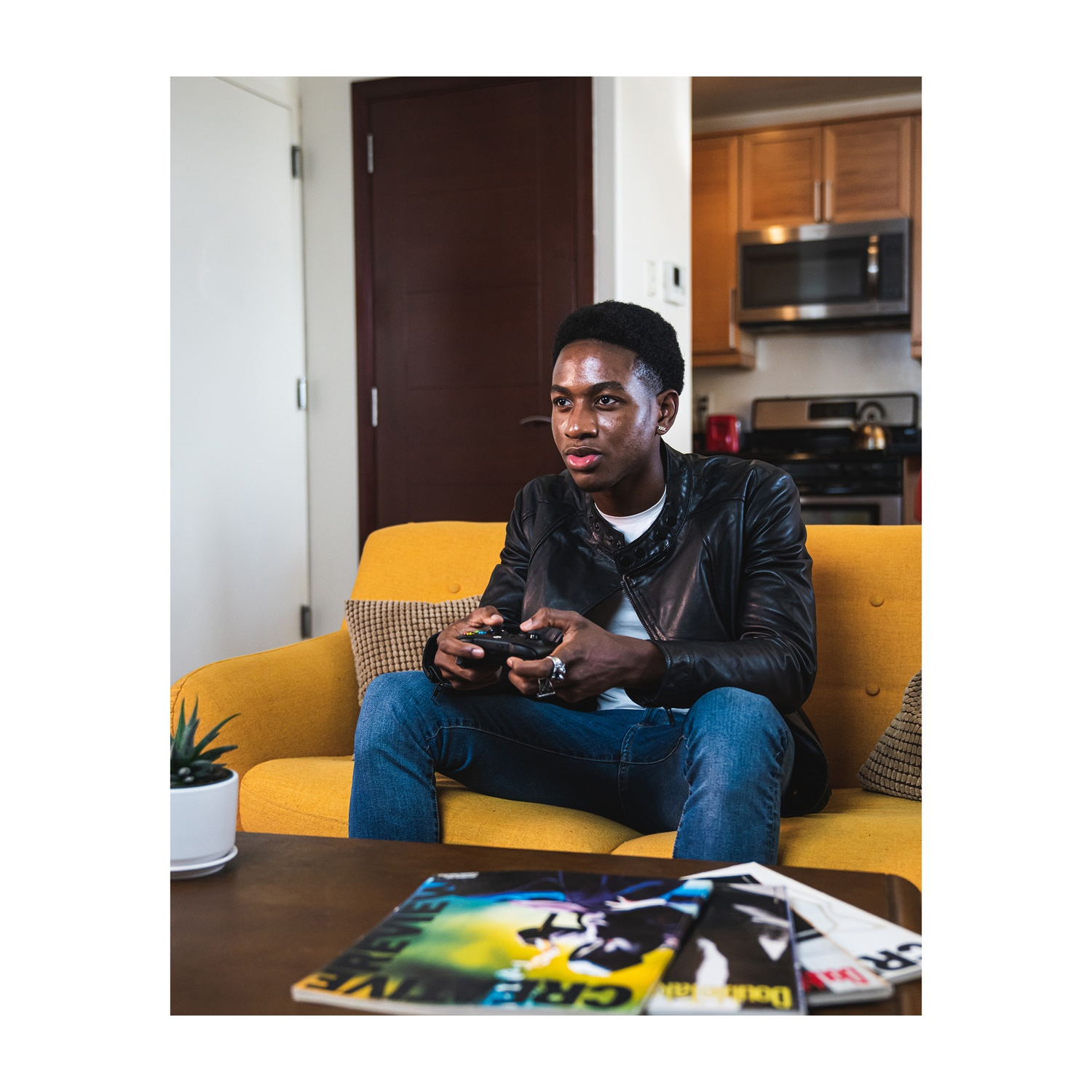 Uomo che gioca ai videogiochi su un divano giallo, tiene un controller. Riviste sul tavolo.