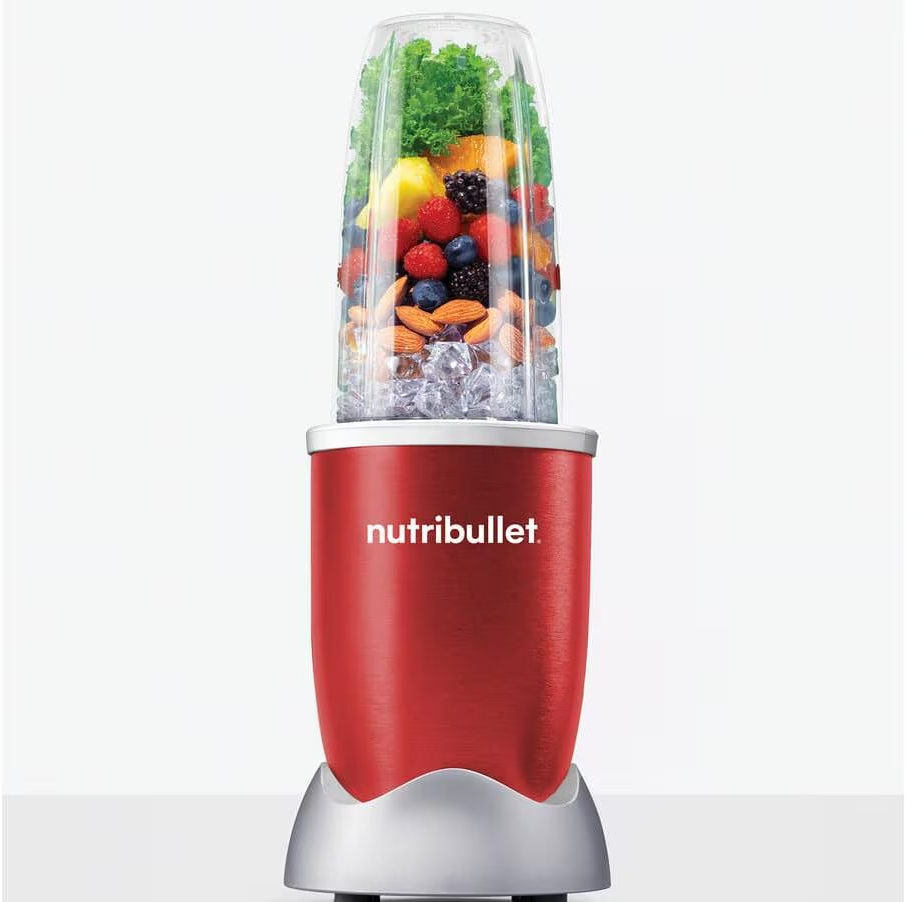 Roter Nutribullet-Mixer gefüllt mit Früchten, Eis und Grünzeug auf weißer Oberfläche.