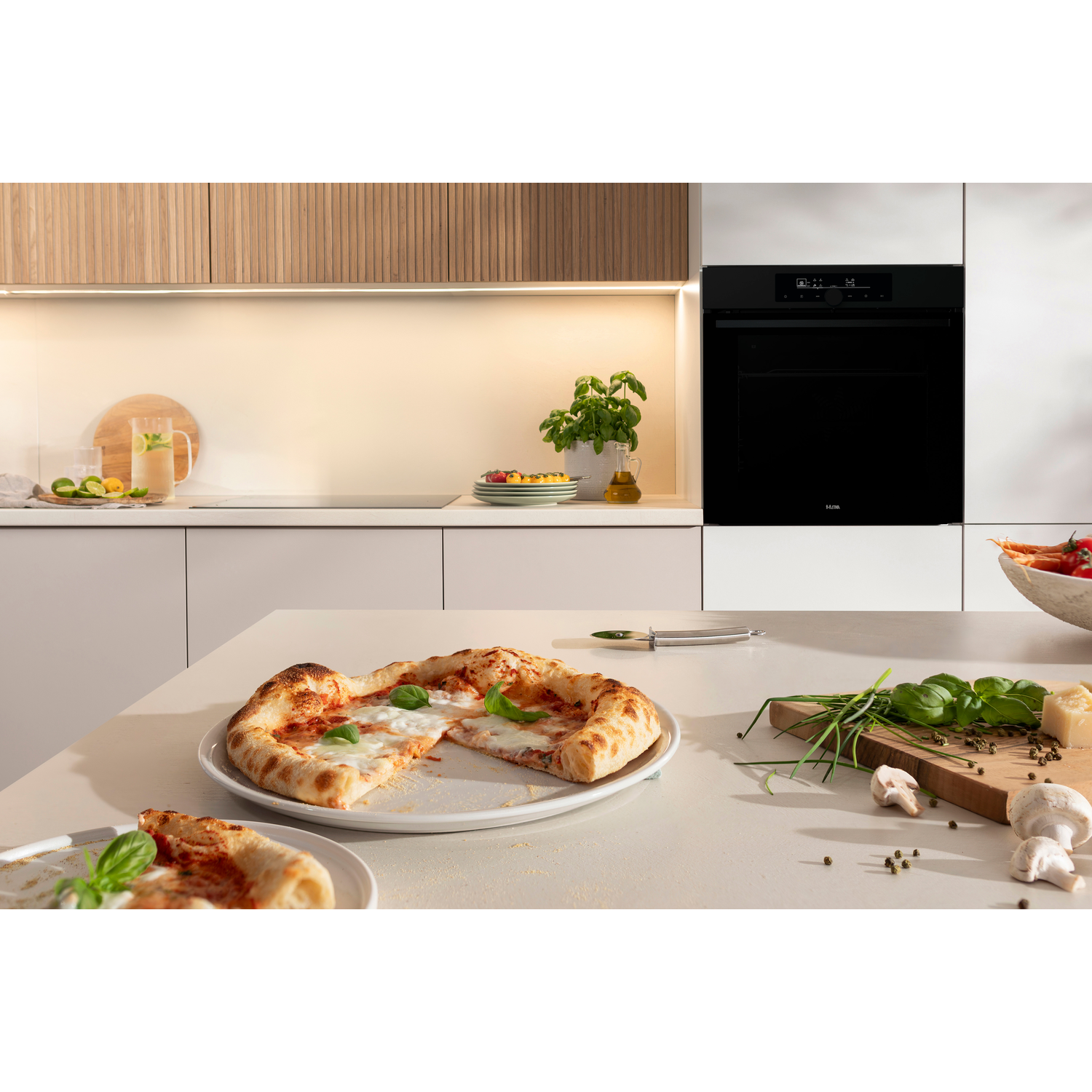 A kitchen scene with a pizza on a table. There is an oven, ingredients, and other food items.
