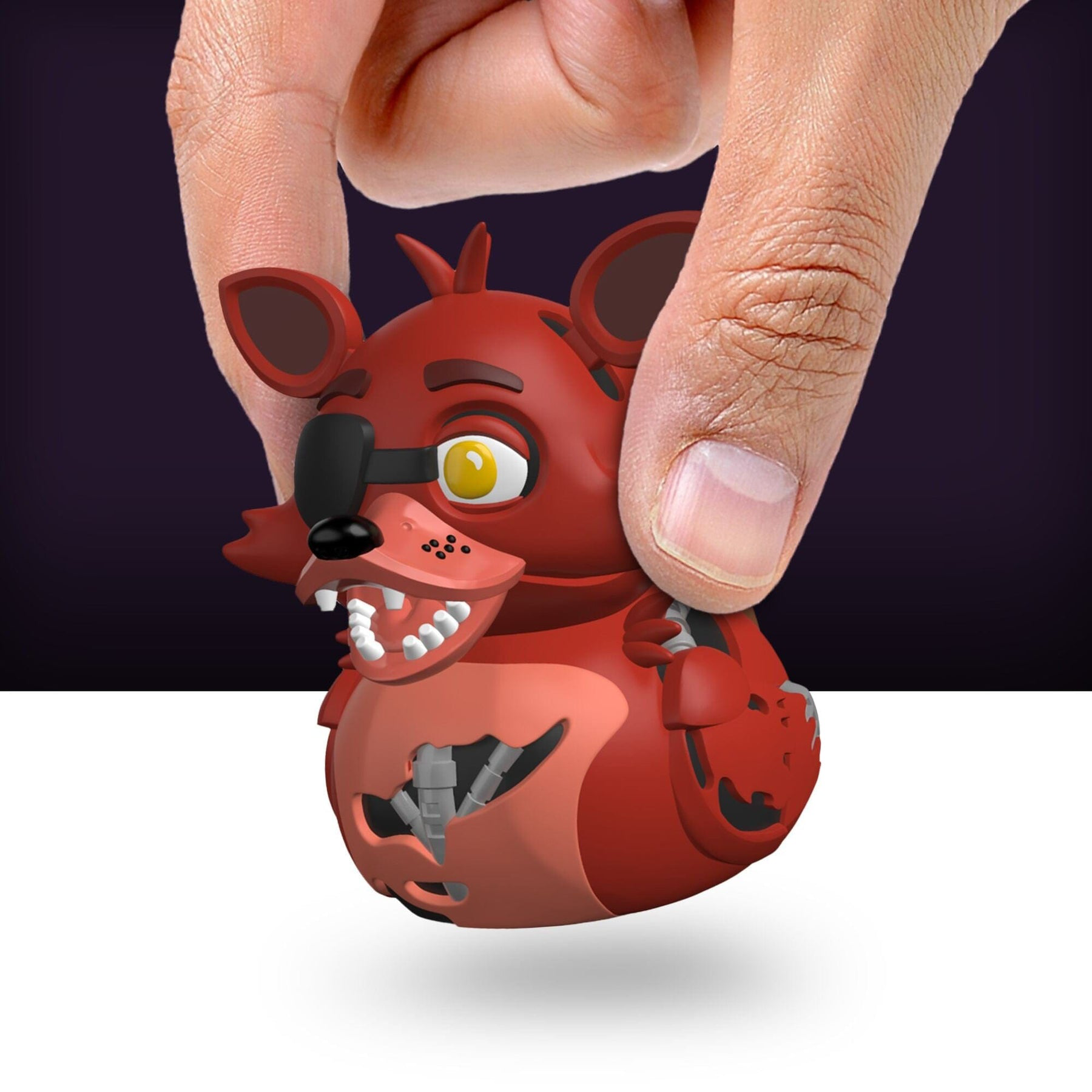 Kéz egy piros Foxy gumikacsát tart. A kacsának szemkötője van. Sötét lila és fehér háttér.