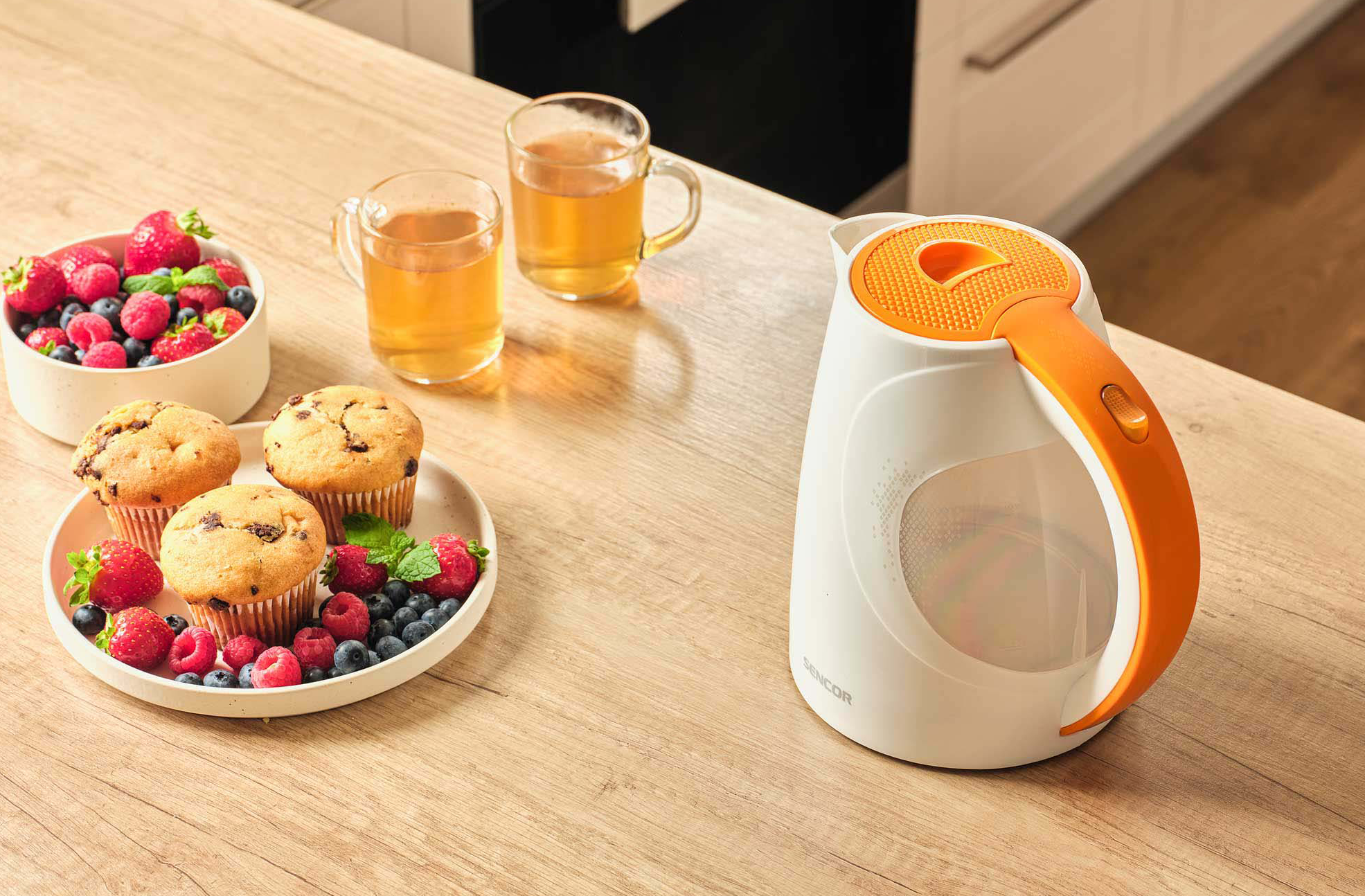 Reggeli jelenet faasztalon: muffinok, bogyók, tea poharakban, fehér-narancs vízforraló.