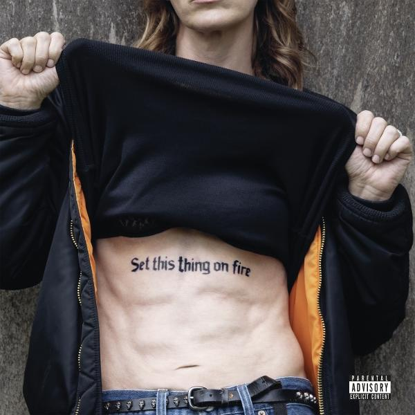 Une personne soulève une veste noire. Un tatouage sur le ventre dit : "Set this thing on fire".