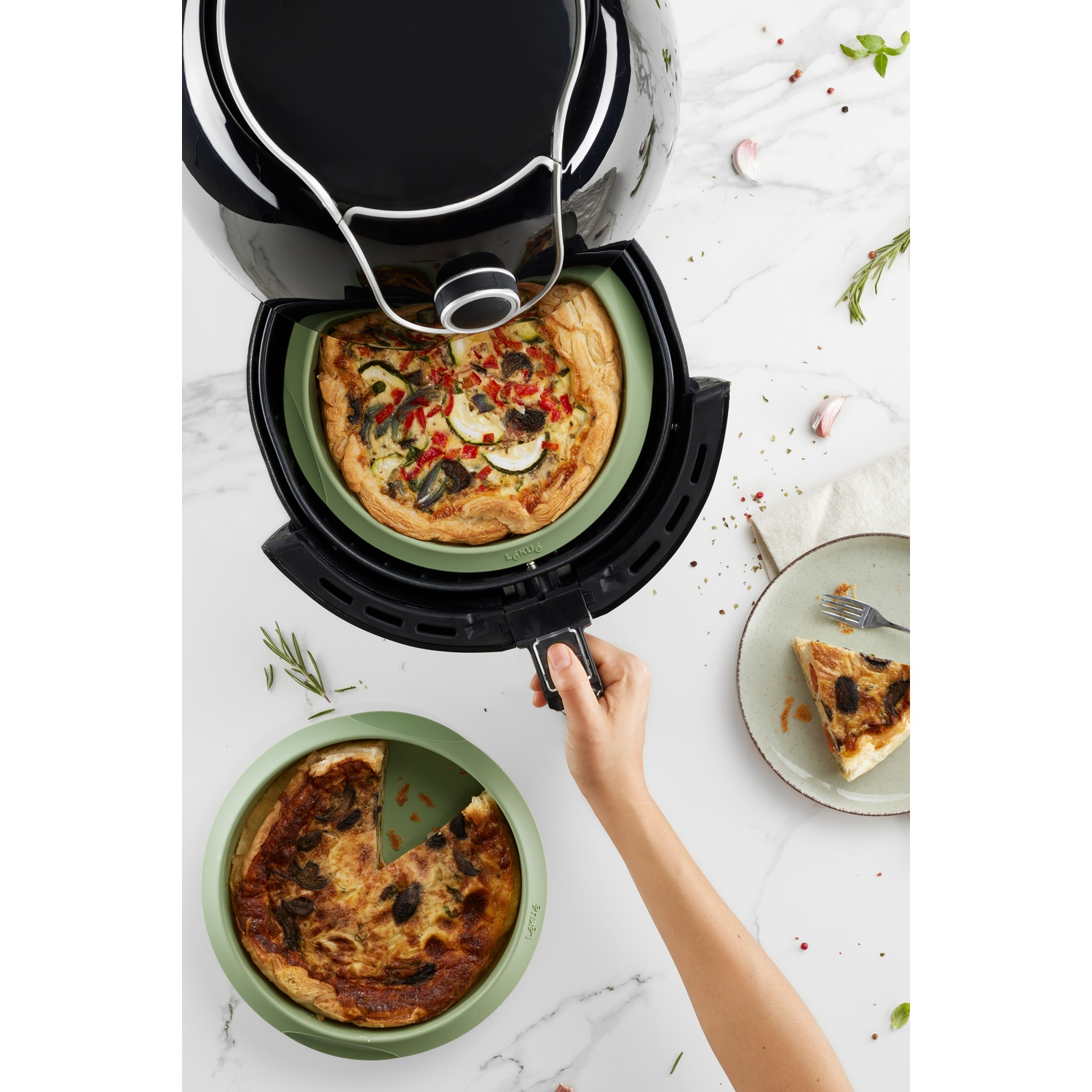 An air fryer with a quiche inside, a slice on a plate, and a second quiche.
