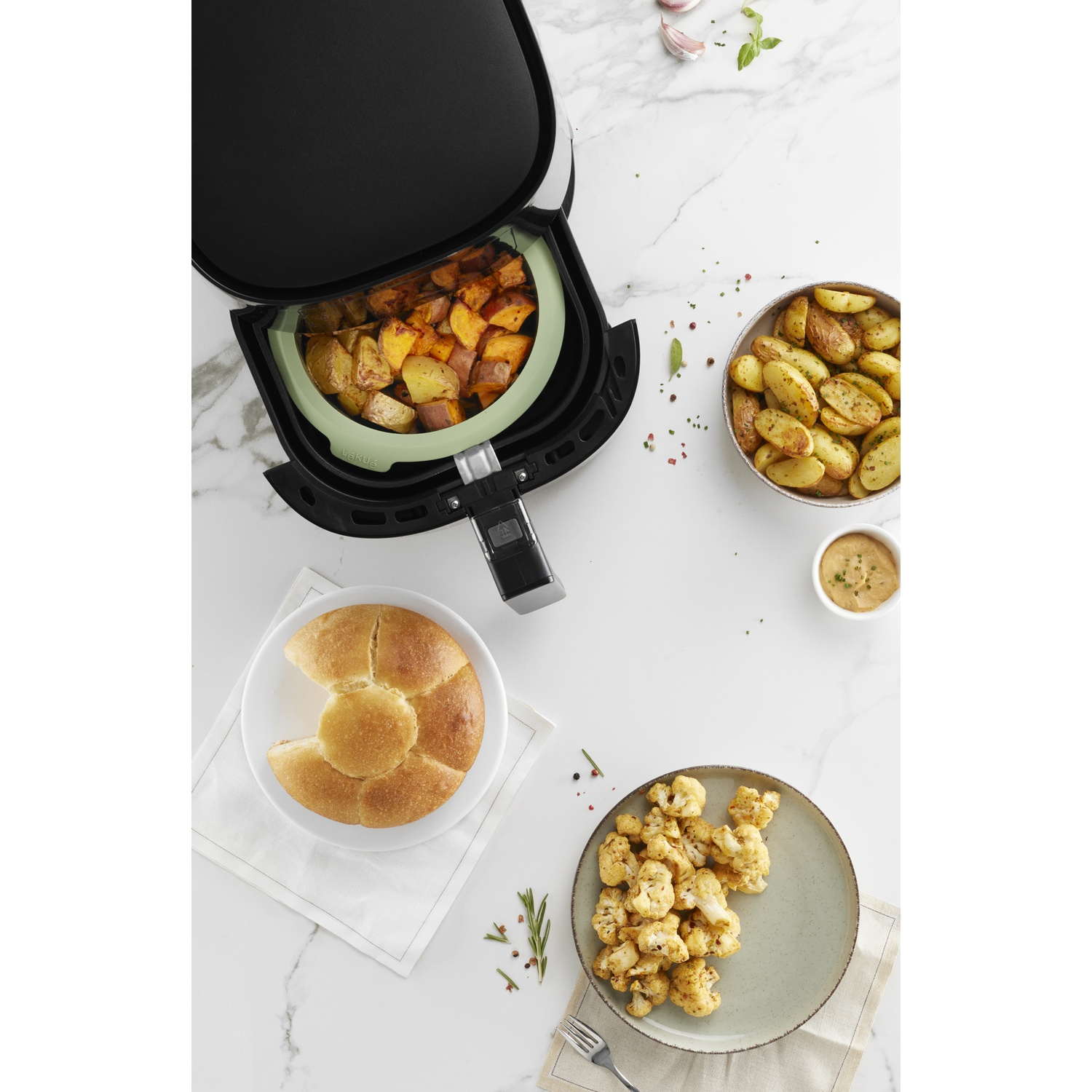 Air fryer with cooked potatoes and vegetables on a white marble surface.
