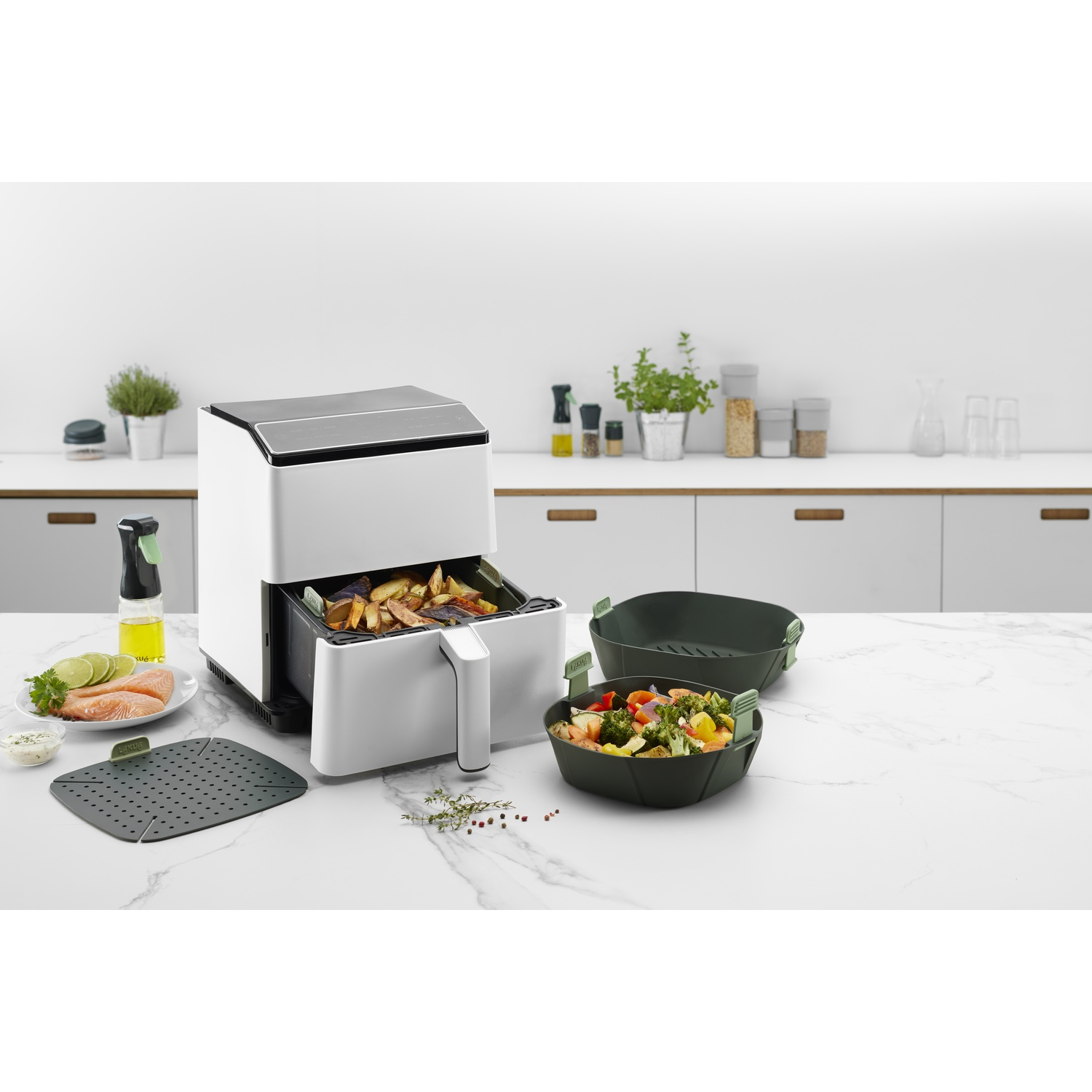 White air fryer with food and accessories. Vegetables, herbs and oil are on the counter.