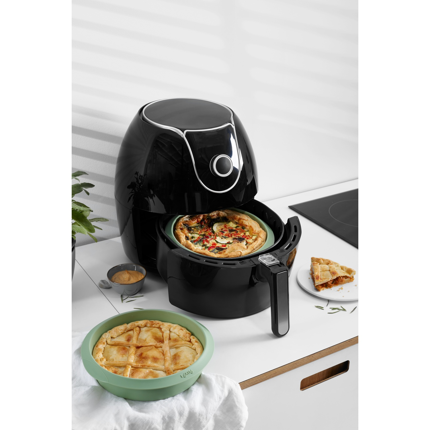 Zwarte airfryer met eten erin. Inclusief pizza en taart. Geplaatst op een wit oppervlak.