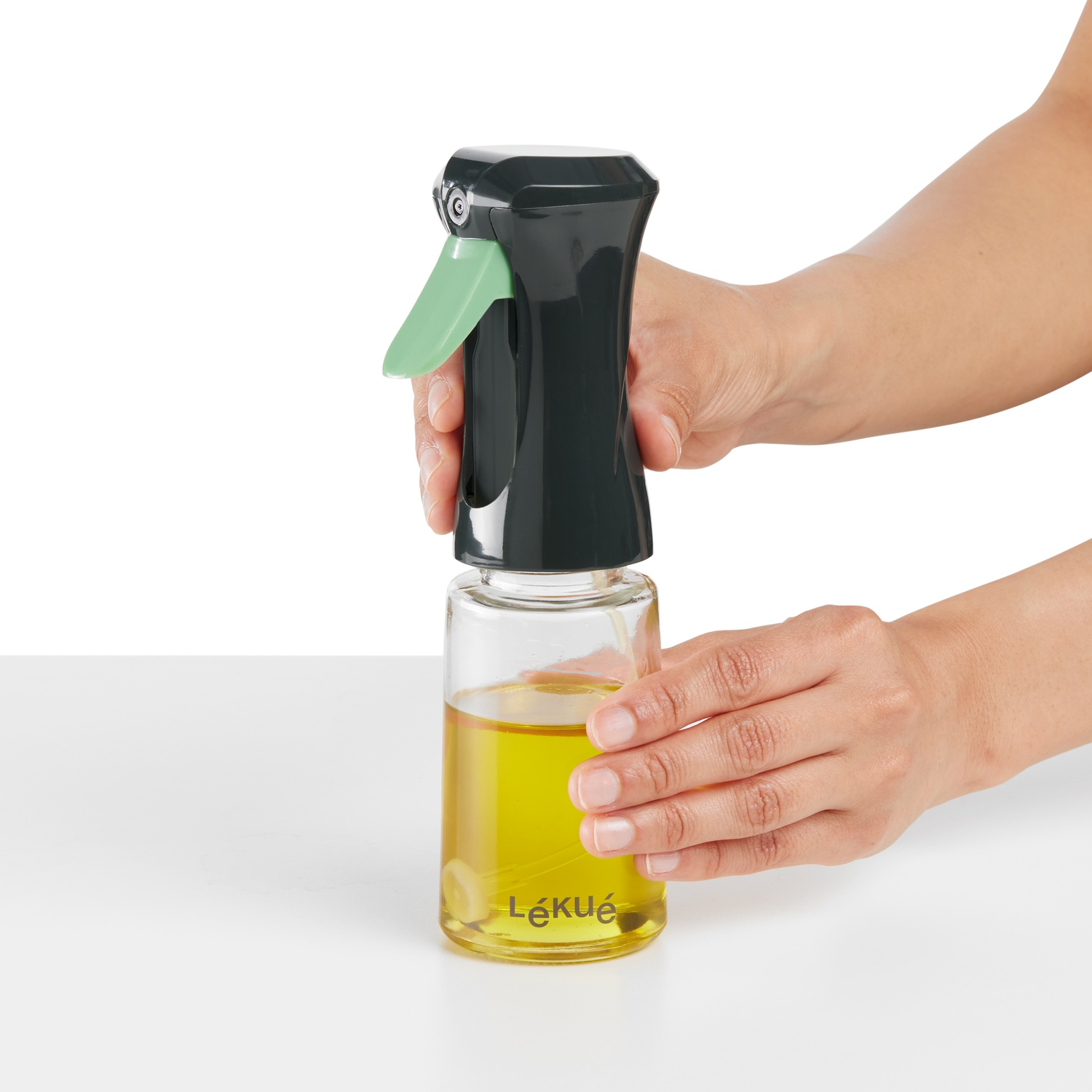 Clear glass oil sprayer with black and green cap, hands holding it.