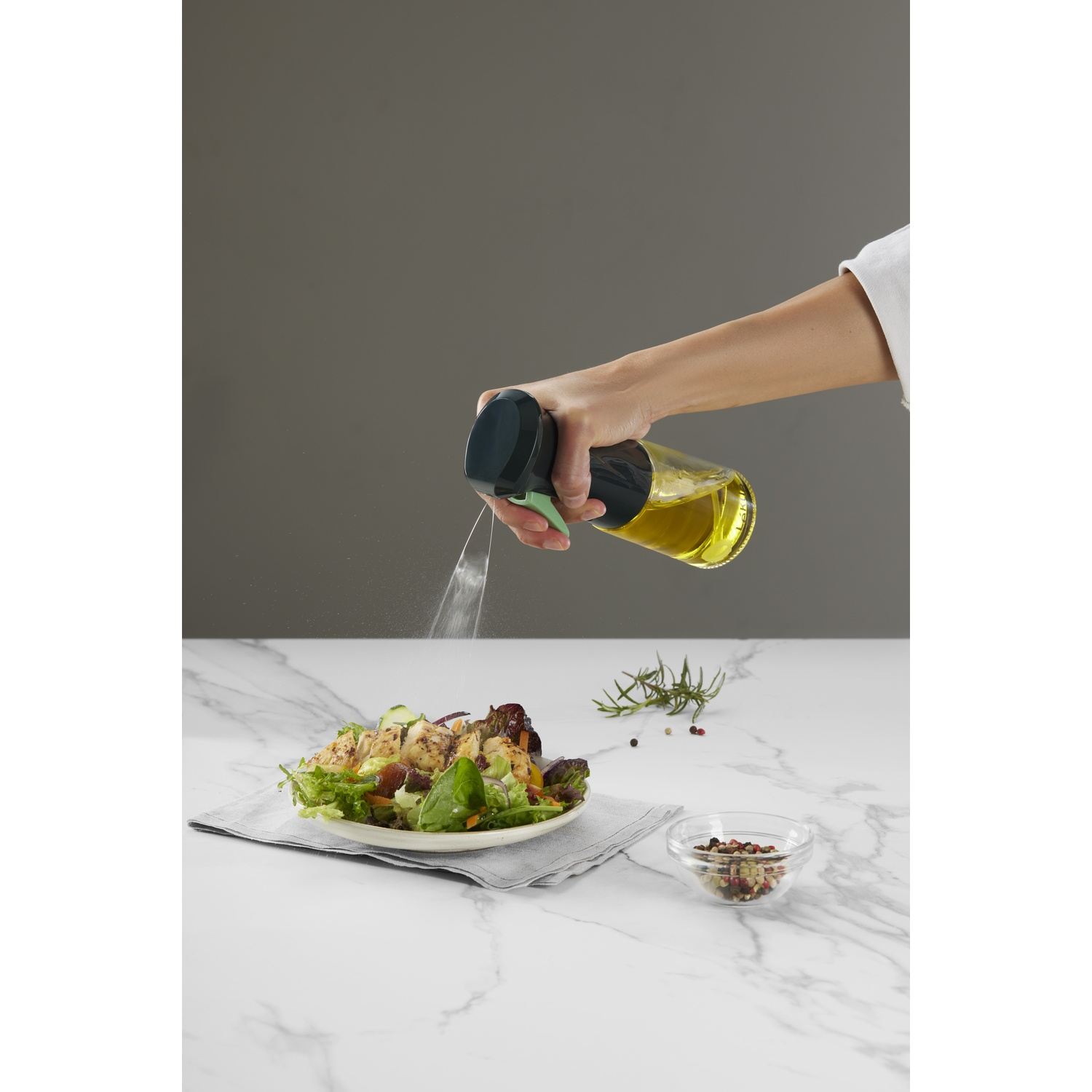 A person spraying oil over a salad in a bowl. A bottle of oil is in their hand.
