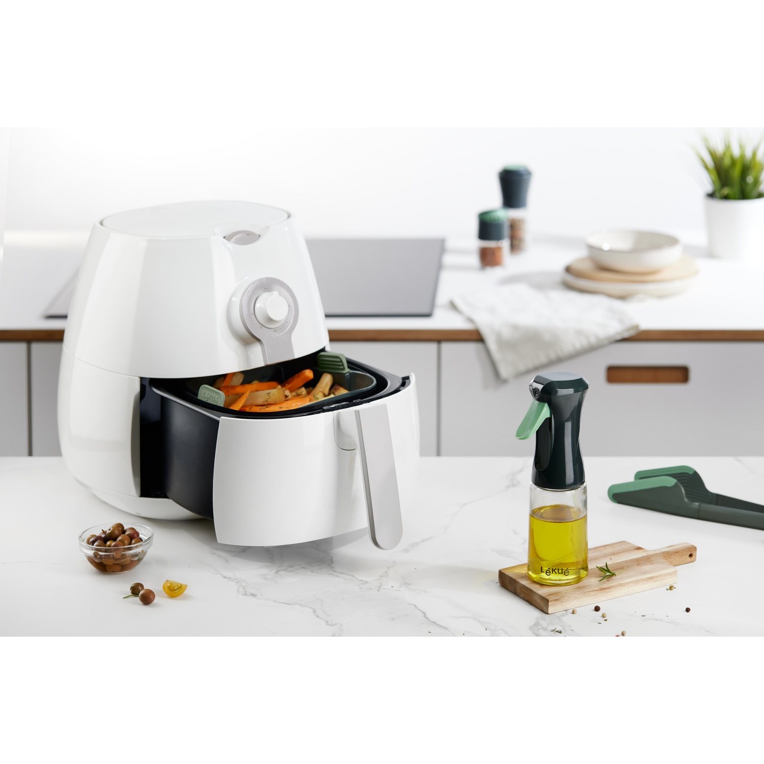 White air fryer with food inside, a bottle of oil, and other kitchen tools on a white marble surface.