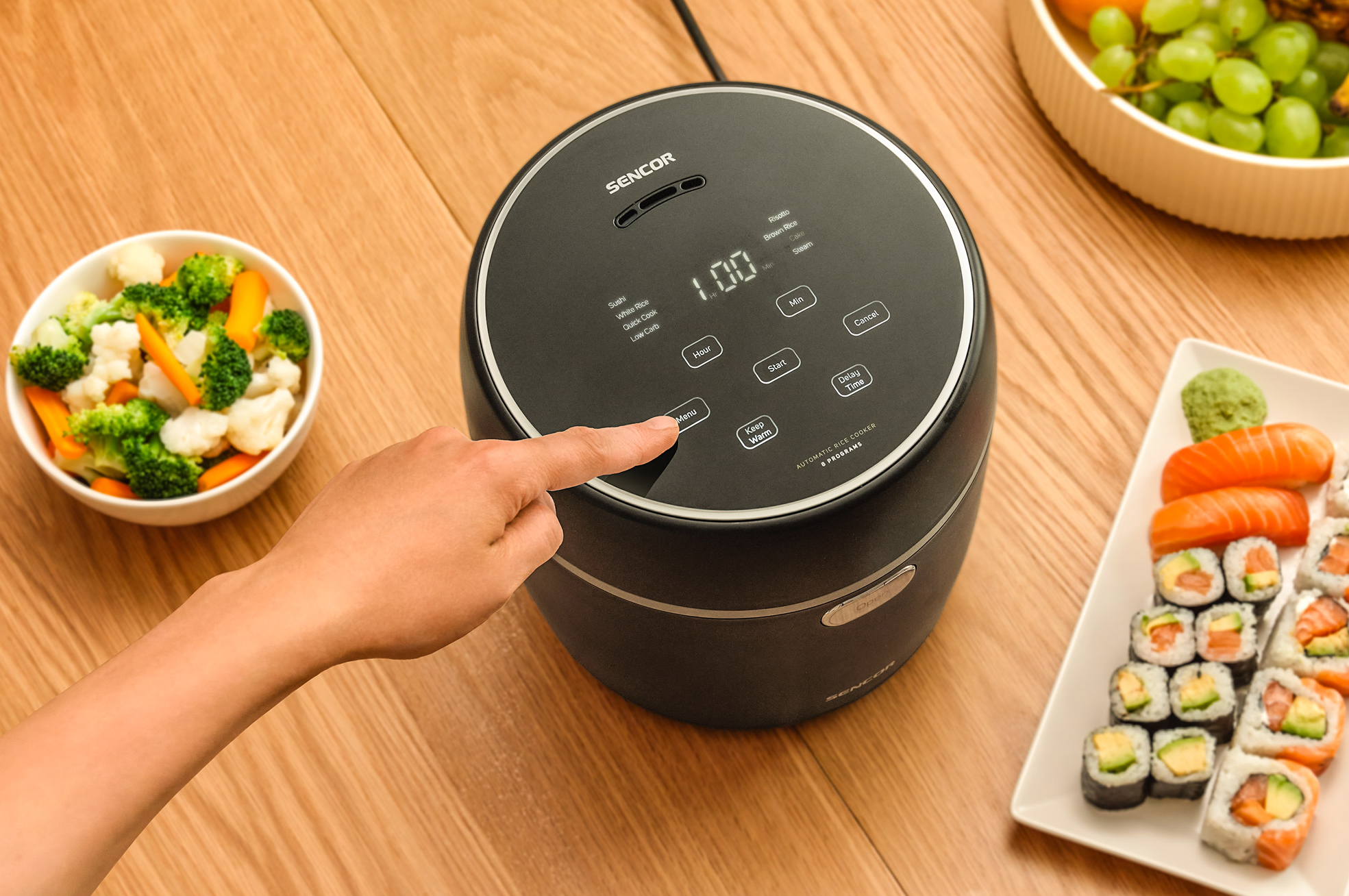 Egy személy keze megnyomja a 'Menü' gombot egy fekete rizsfőzőn. Sushi és zöldségek az asztalon.