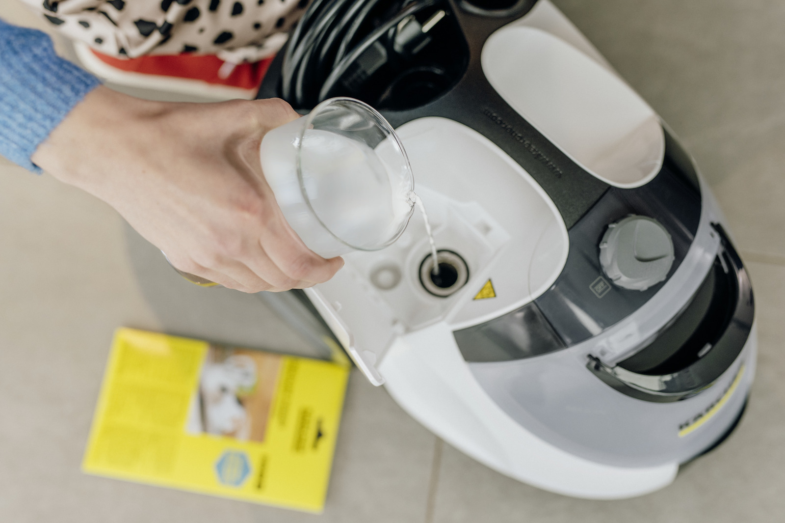 Osoba wlewa wodę do białej myjki parowej. Myjka jest szara i czarna.