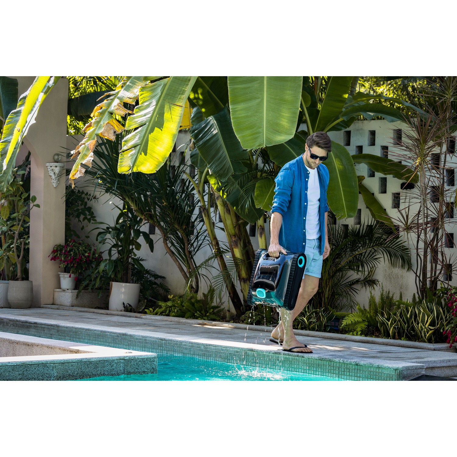 Un uomo cammina a bordo piscina tenendo in mano un pulitore per piscina. Sullo sfondo palme e piante.