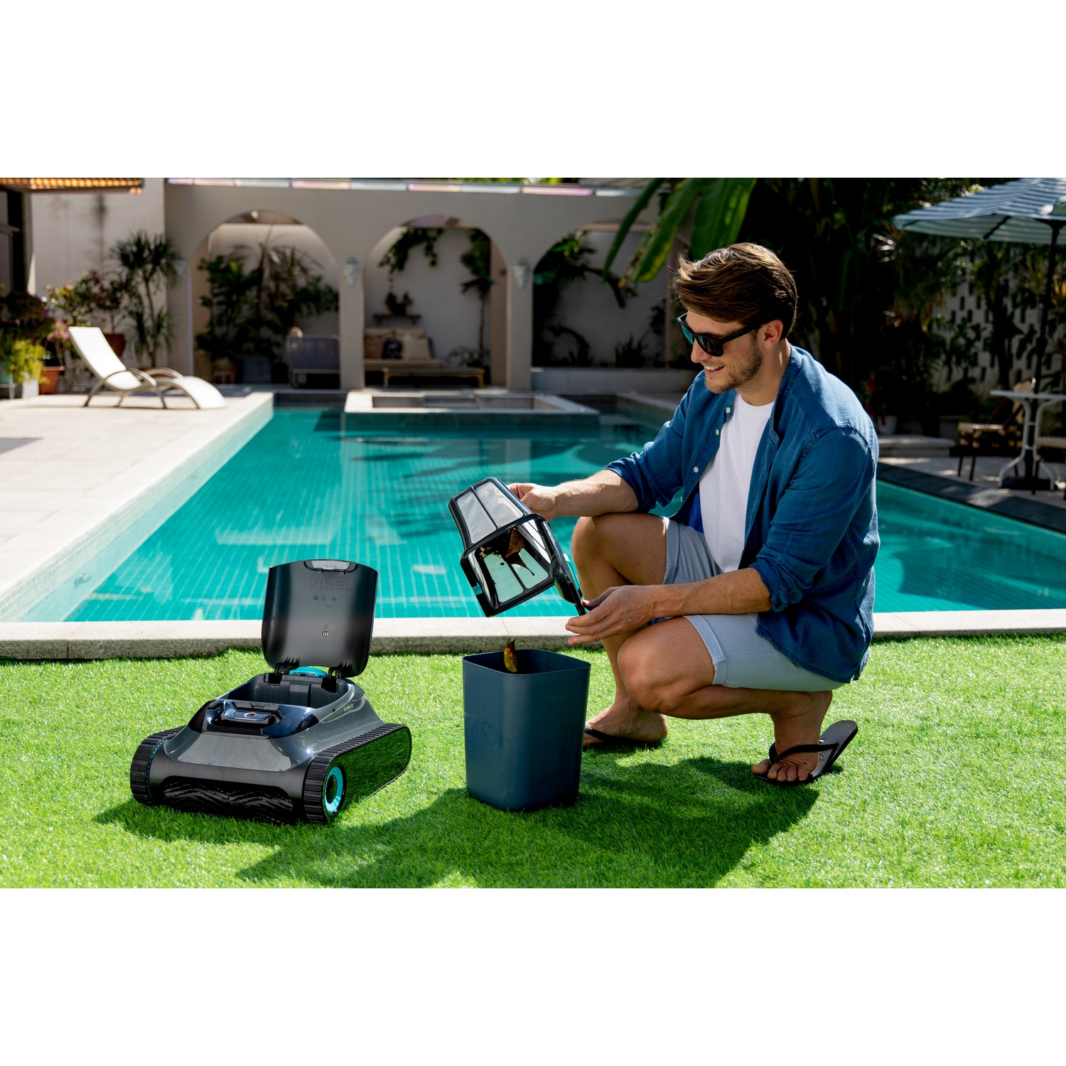 Un uomo svuota il filtro di un robot pulitore per piscina in un bidone accanto a una piscina.