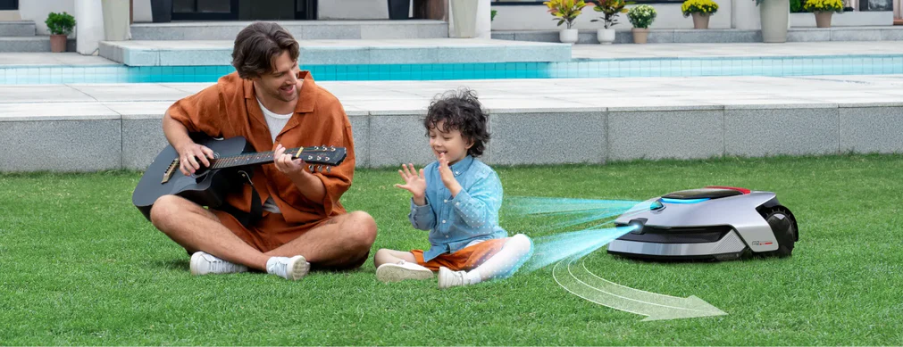 Ein Mann spielt Gitarre für ein Kind, neben einem Roboter-Rasenmäher auf grünem Gras. Im Hintergrund sind ein Pool und ein Haus.