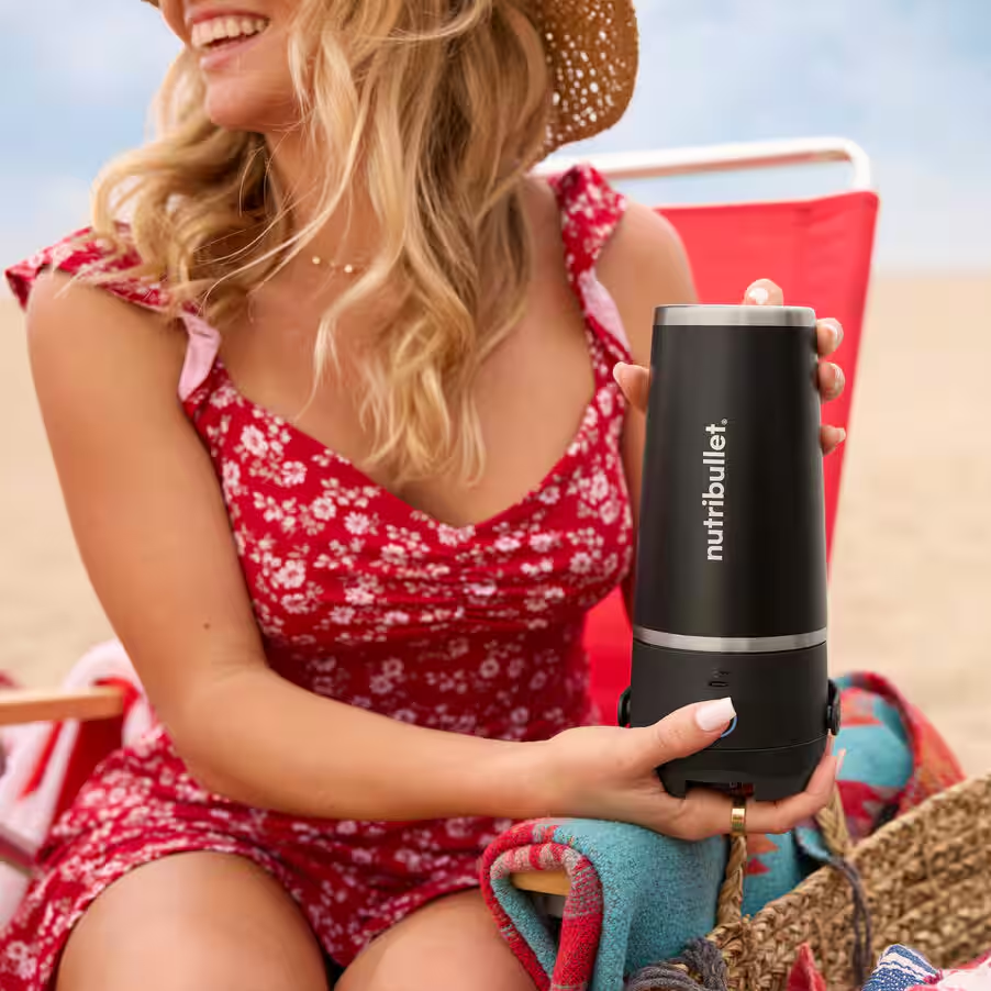 Una donna con un vestito rosso tiene in mano un Nutribullet nero, con la spiaggia sullo sfondo.