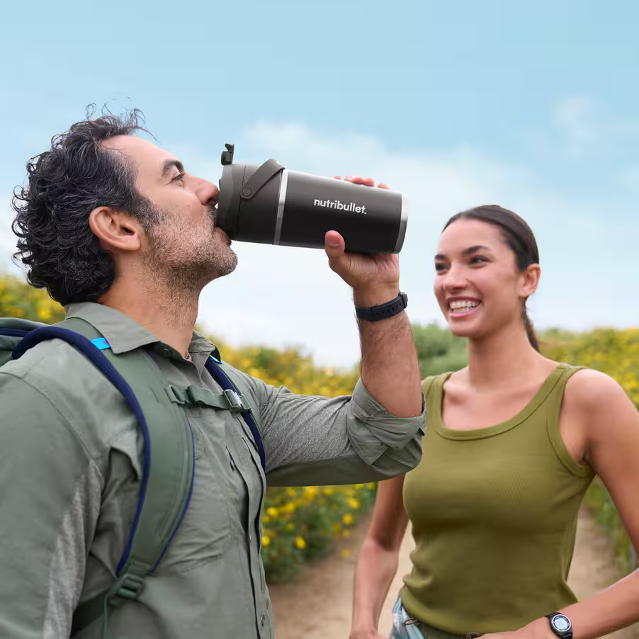Uomo beve da borraccia nera, donna sorride sullo sfondo, ambiente esterno.