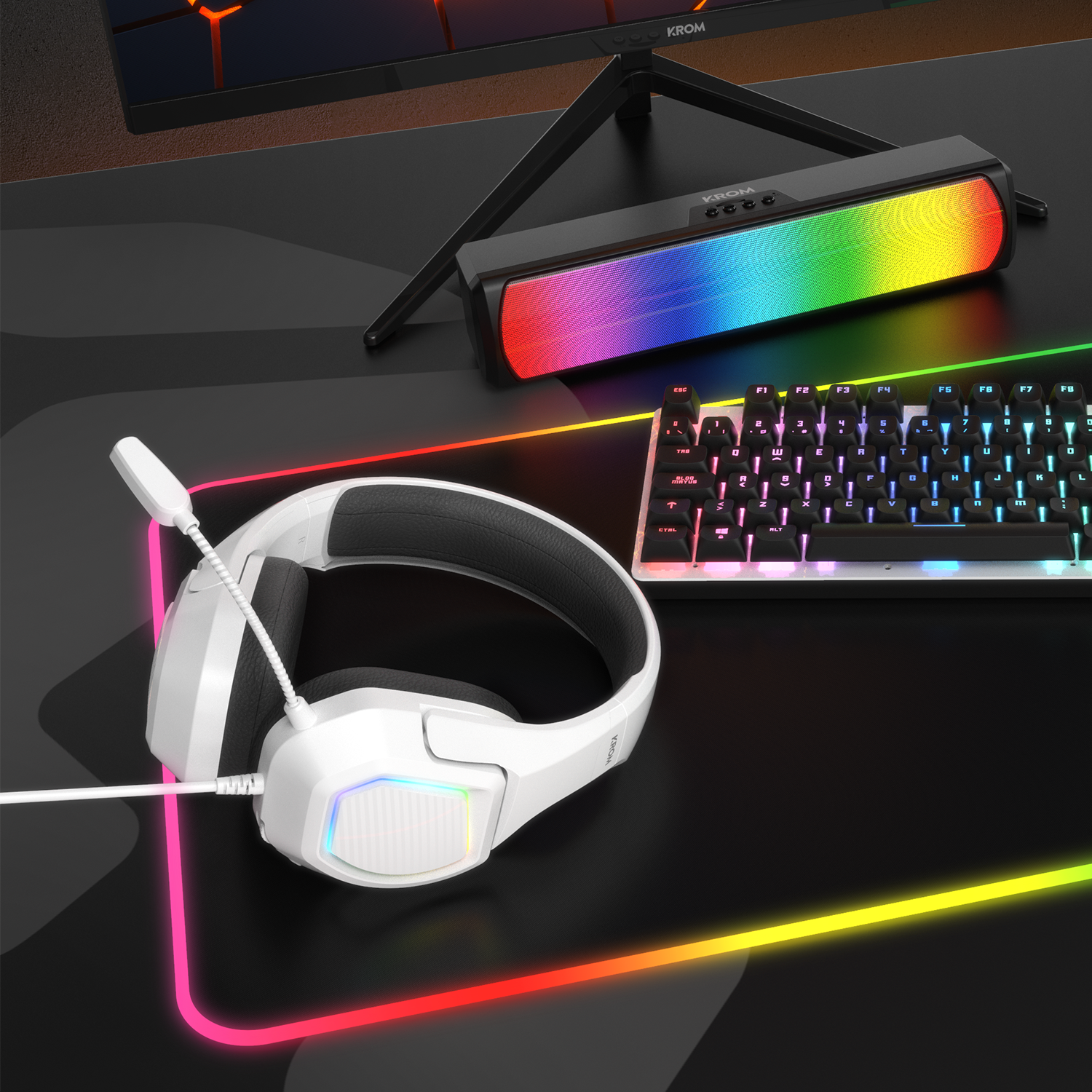 Configuración gaming con altavoz KROM, teclado, auriculares y alfombrilla con bordes brillantes.