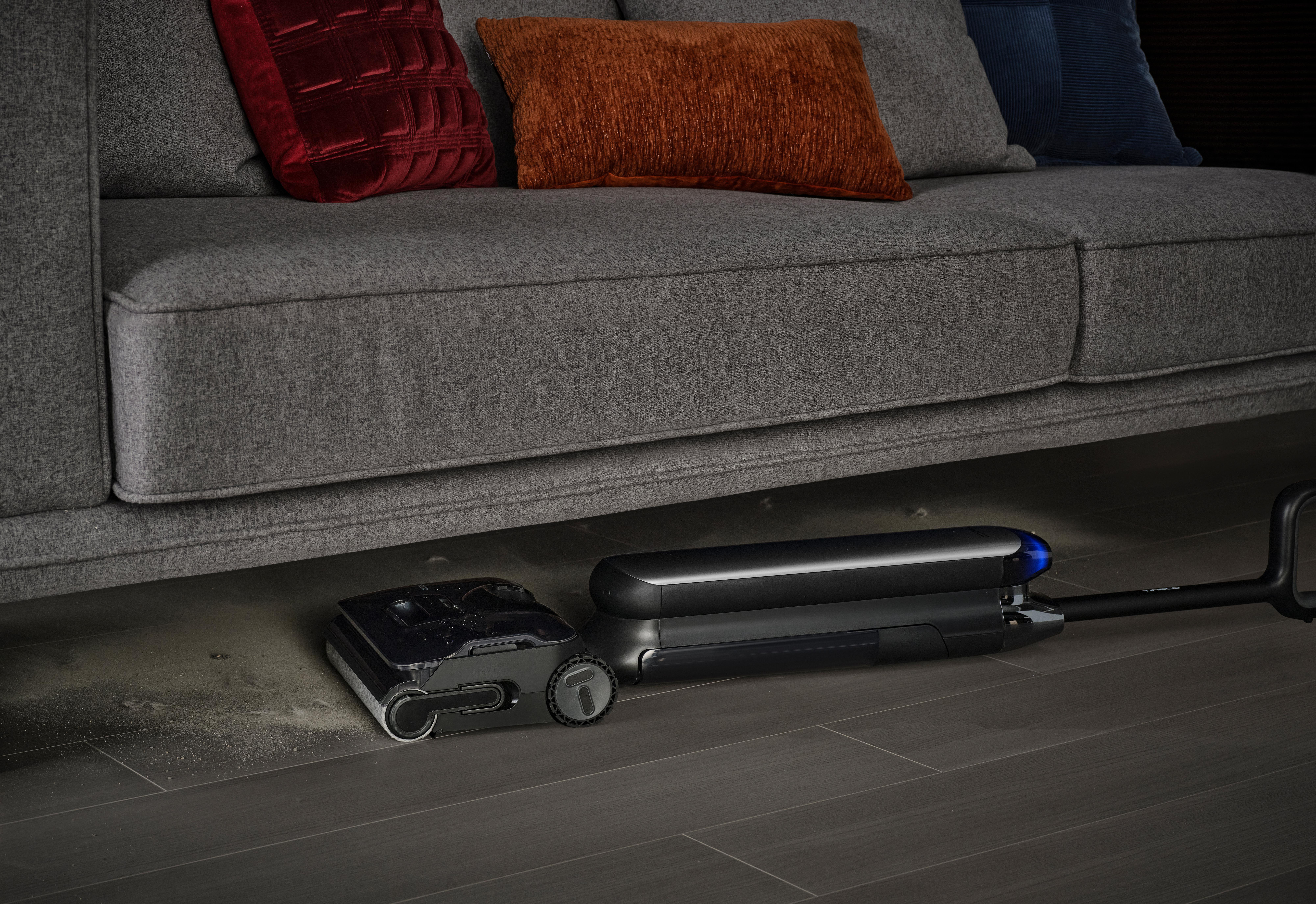 Ein Roboter-Staubsauger reinigt unter einer grauen Couch auf einem Holzboden.