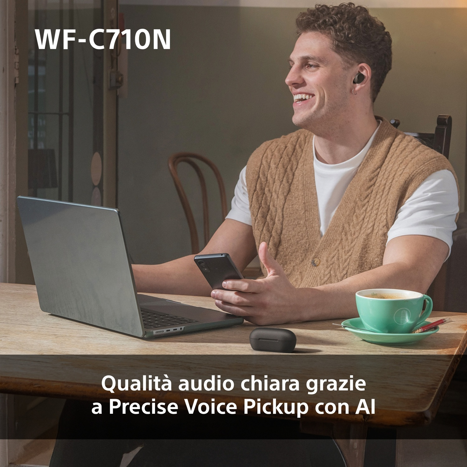 Uomo sorride, con maglione, usa laptop, telefono e auricolari. Tazza verde sul tavolo.