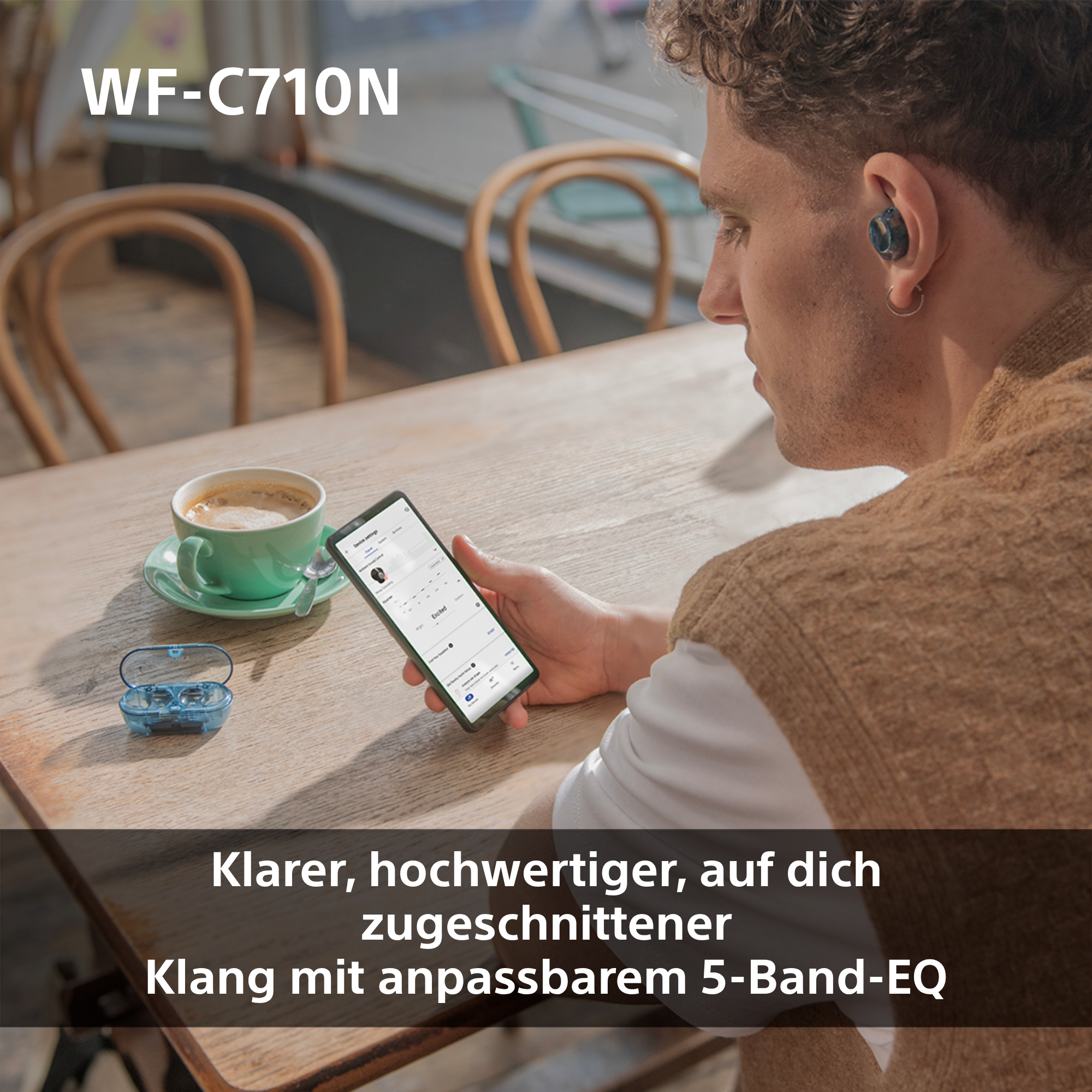 Ein Mann mit Ohrhörern schaut auf ein Telefon. Auf dem Tisch stehen Kaffee und eine Ohrhörer-Hülle.
