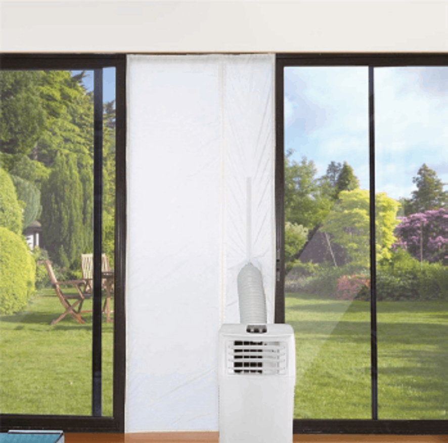 Un acondicionador de aire portátil frente a una ventana con una cortina blanca. Se ve un jardín a través de la ventana.