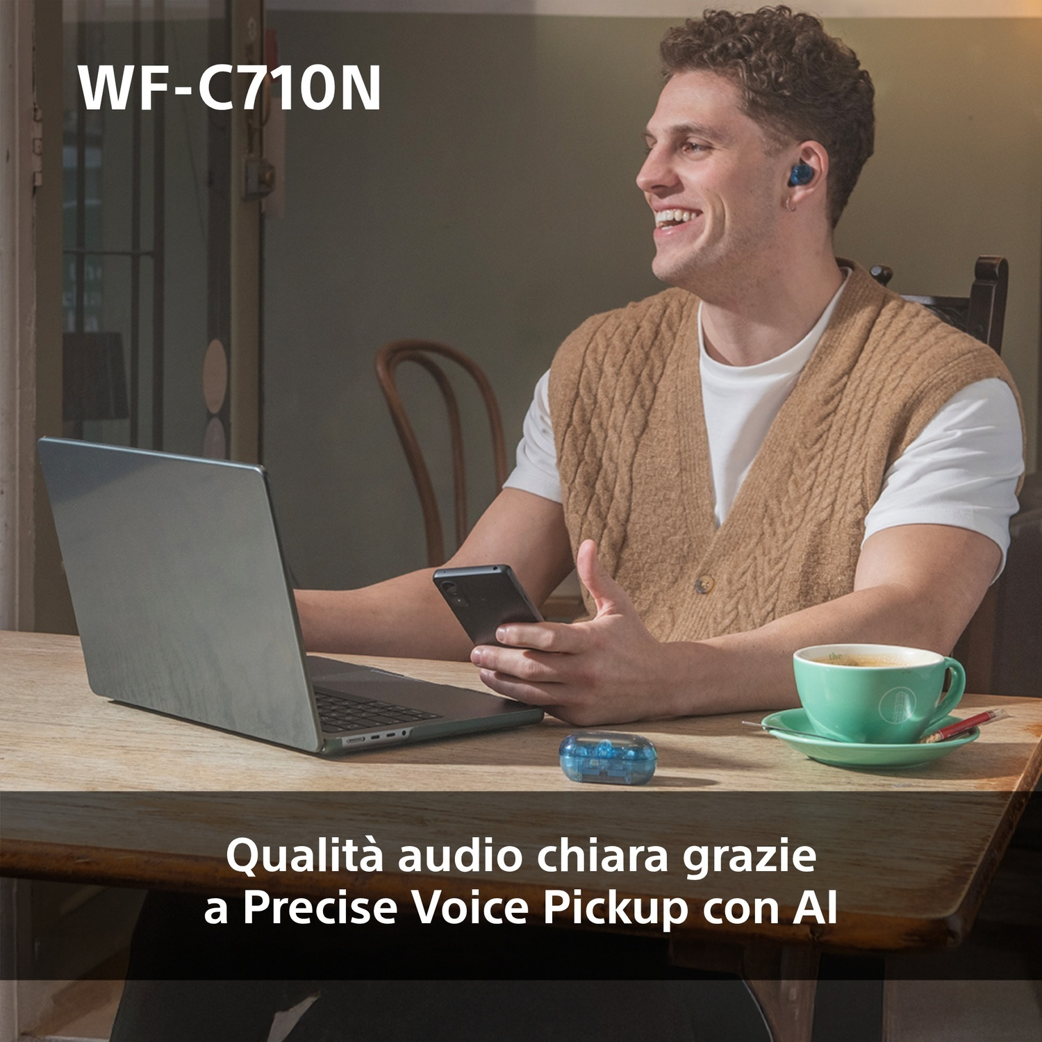 Un uomo con le cuffie sorride usando un laptop. Una tazza di caffè e un telefono sul tavolo.
