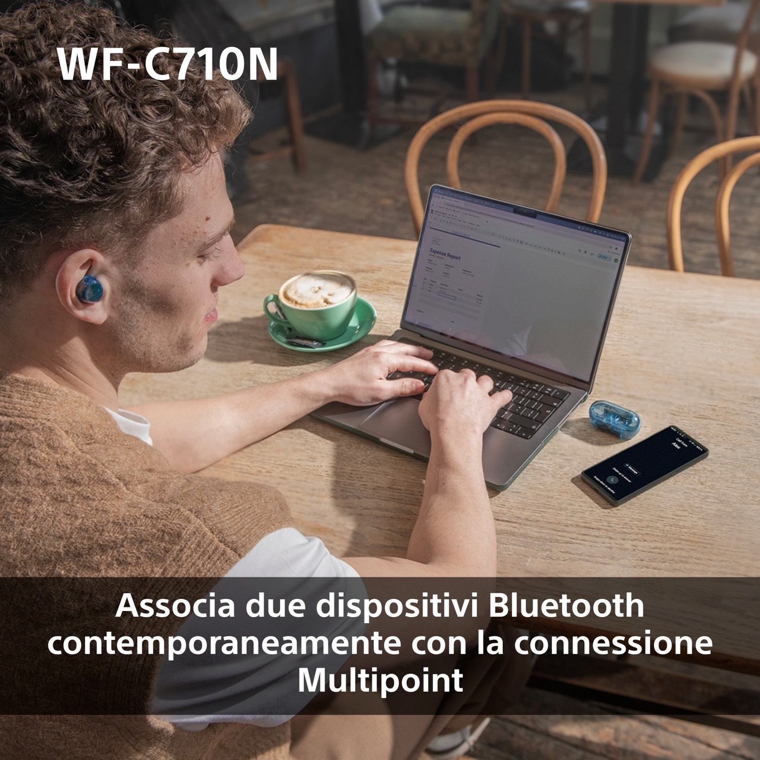 Uomo usa laptop con auricolari e telefono sul tavolo, tazza di caffè, tavolo di legno e sfondo sfocato.