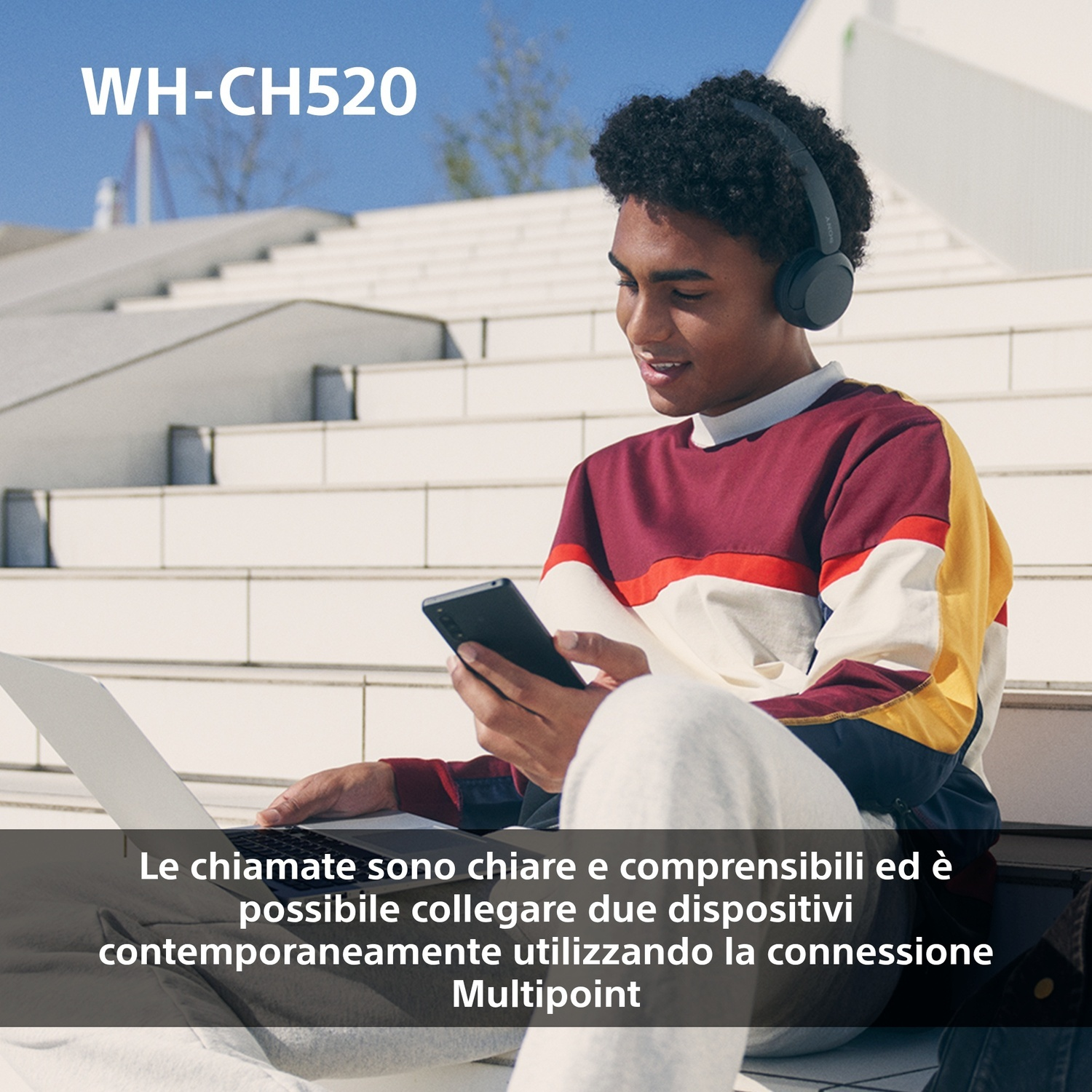 Un giovane con le cuffie usa uno smartphone e un laptop su una scalinata.