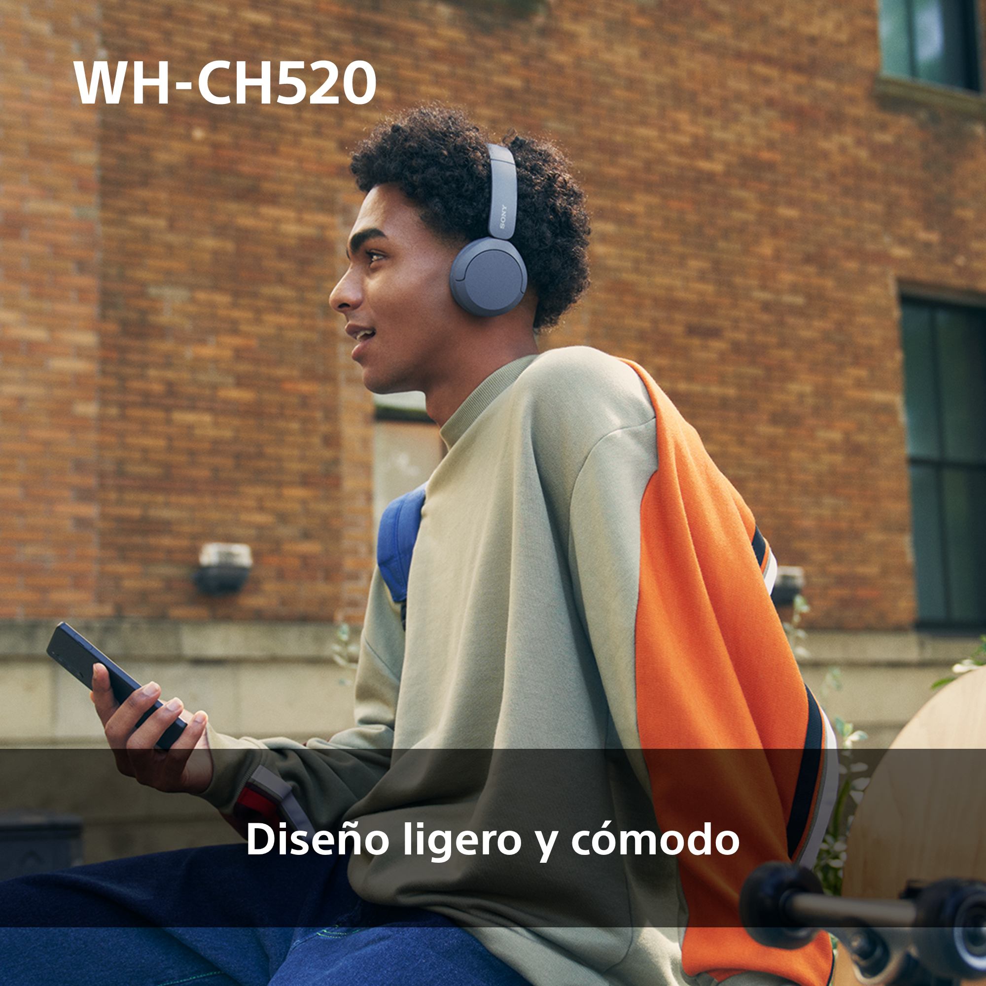 Hombre con auriculares, sosteniendo un teléfono, caminando frente a un edificio de ladrillos.