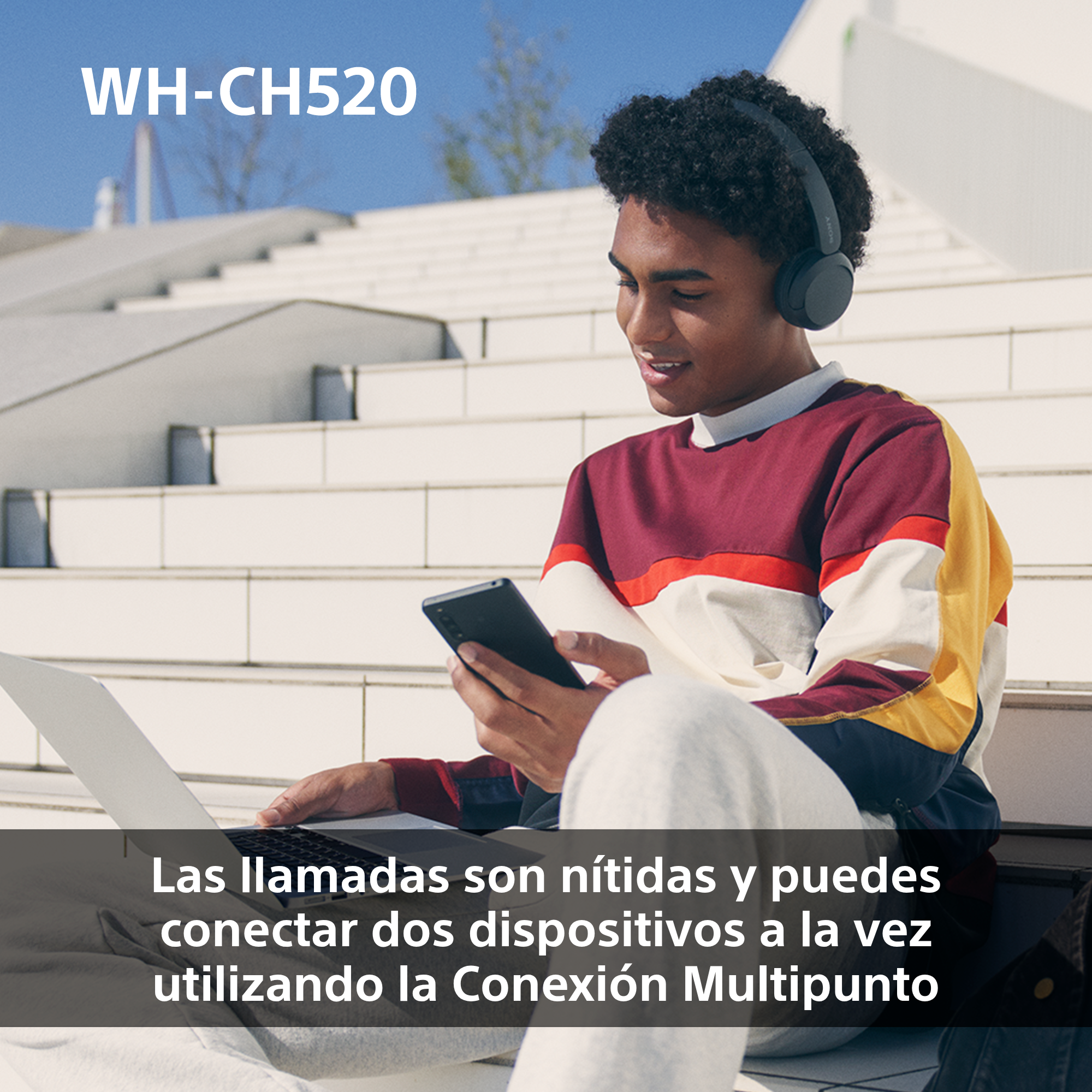 Joven con auriculares y portátil, sonriendo, al aire libre. Teléfono negro en mano.