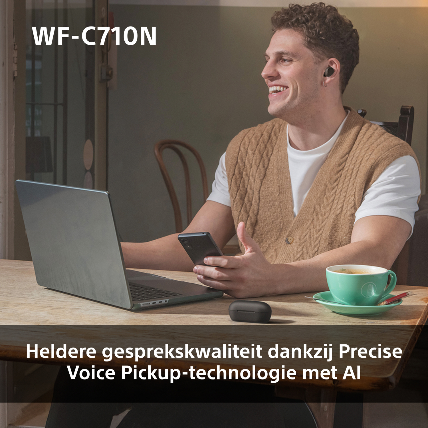 Een man met zwarte oordopjes lacht terwijl hij aan een tafel zit met een laptop en een kop koffie.