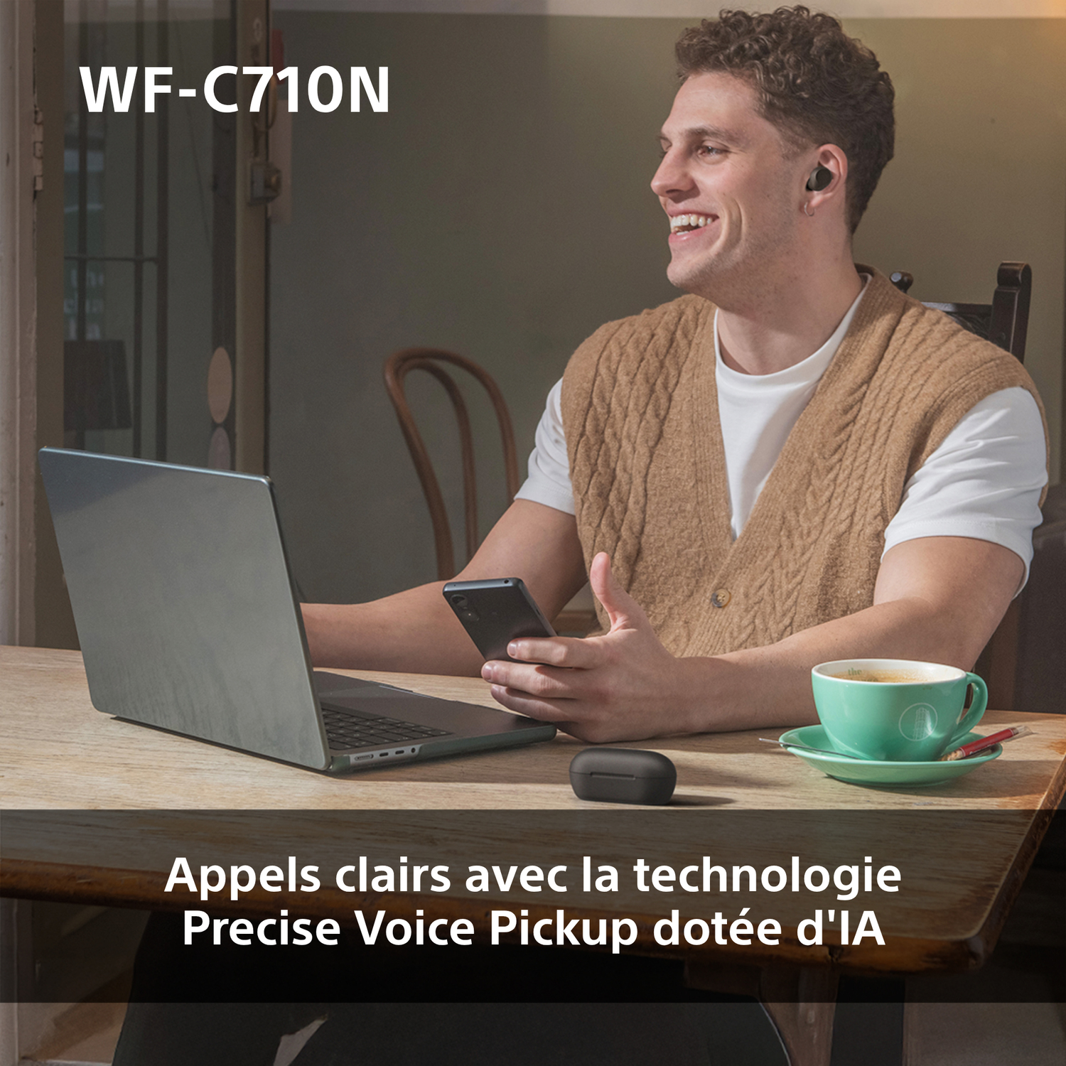 Homme avec écouteurs souriant, tenant un téléphone. Un ordinateur portable, une tasse et un étui pour écouteurs sont sur une table.
