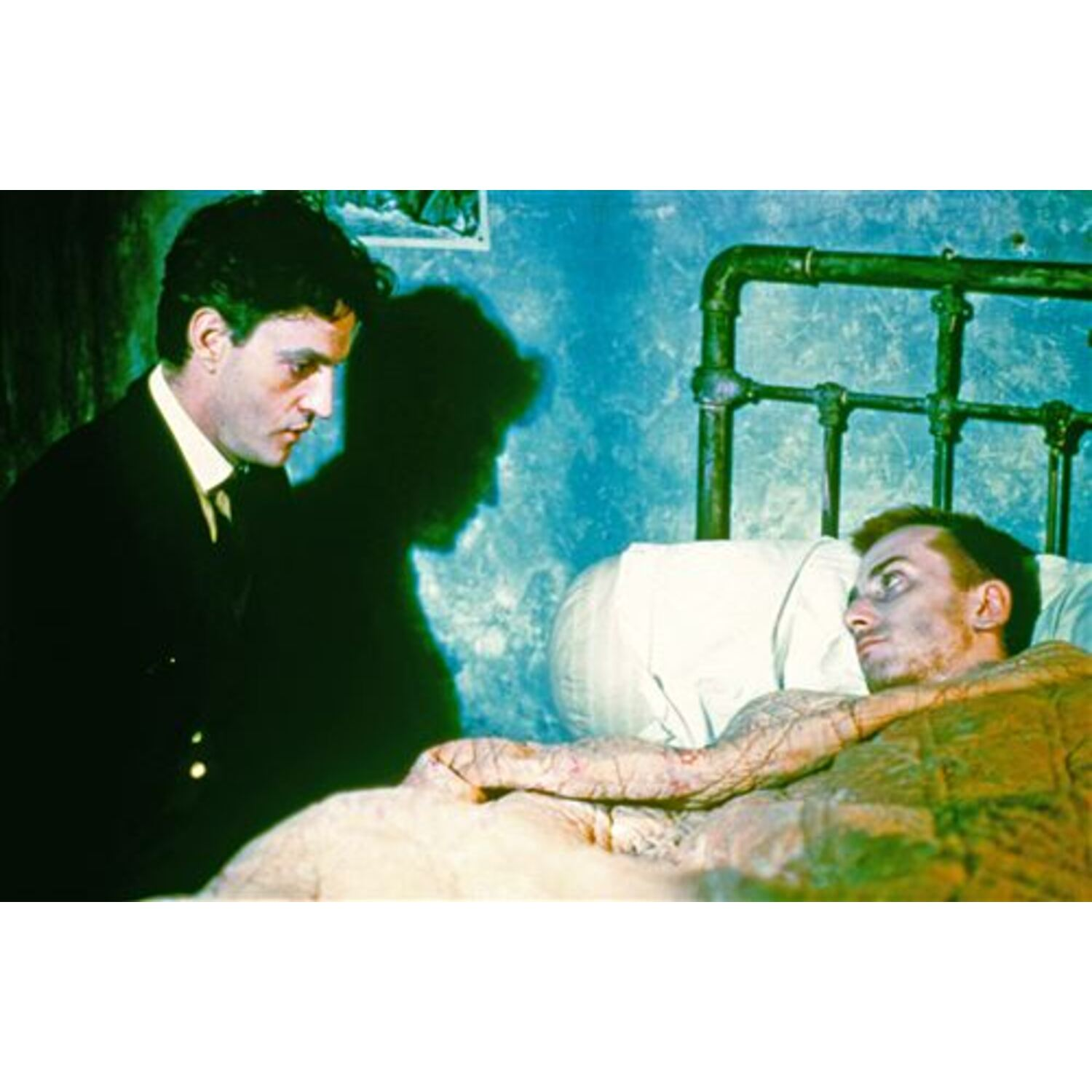 Twee mannen in een kamer, een kijkt naar een ander die in bed ligt, dramatische belichting.