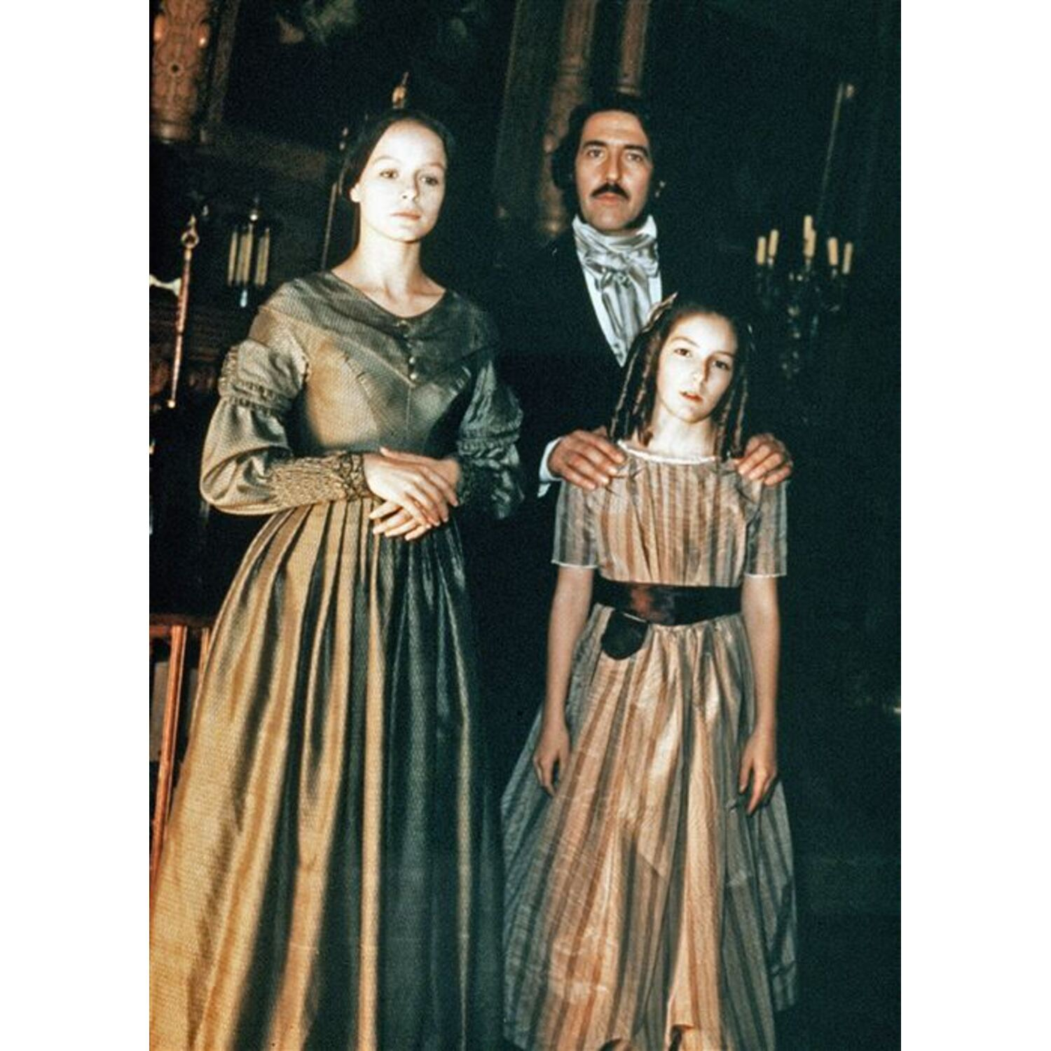 Een familieportret: een vrouw, een man en een meisje staan samen, gekleed in kleding uit die tijd, in een donkere kamer.