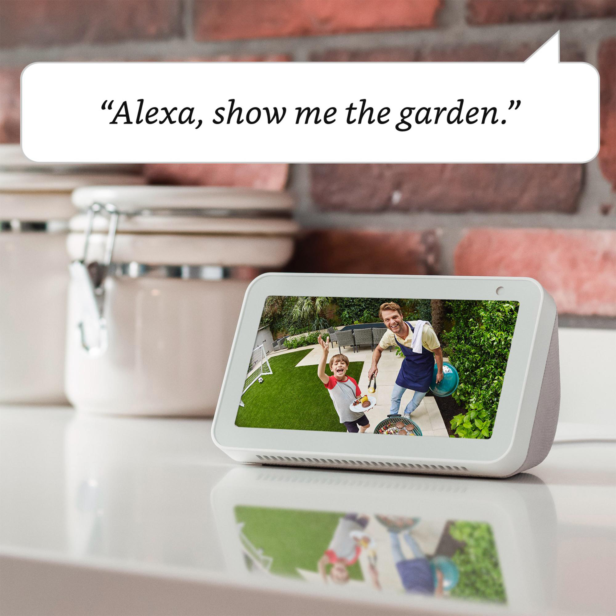 Smart Display zeigt Garten-Video. Auf weißer Oberfläche, Ziegelwandhintergrund. Alexa-Sprachaufforderung.