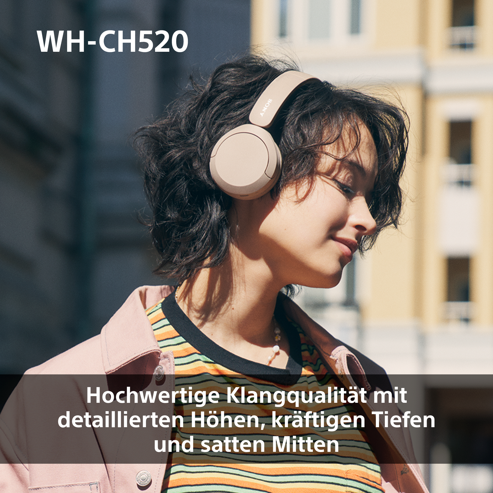 Eine Frau mit beigen Kopfhörern lächelt. Sie trägt ein gestreiftes Oberteil und eine rosa Jacke. Der Hintergrund ist unscharf.