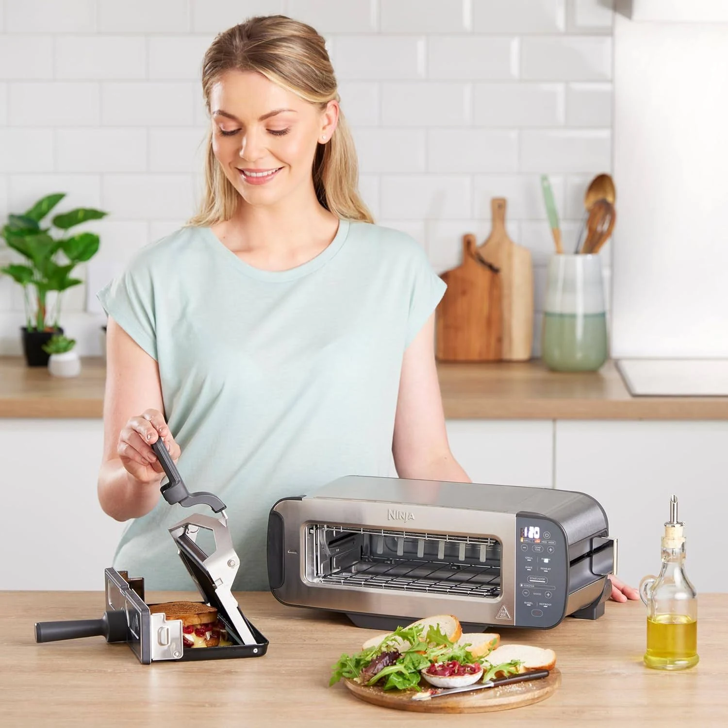 Mujer en una cocina usando una sandwichera. Una tostadora, un sándwich y aceite están en la mesa. Una planta y tablas de cortar están en el fondo.