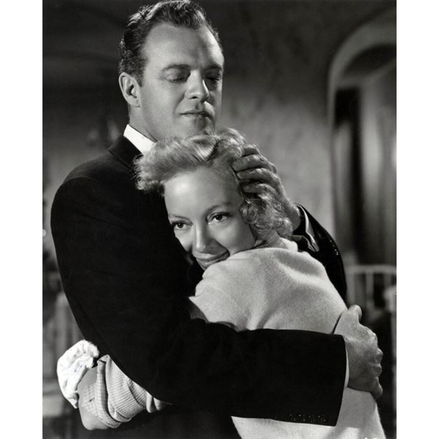Un homme en costume noir enlace une femme en pull clair, intérieur, image sépia.