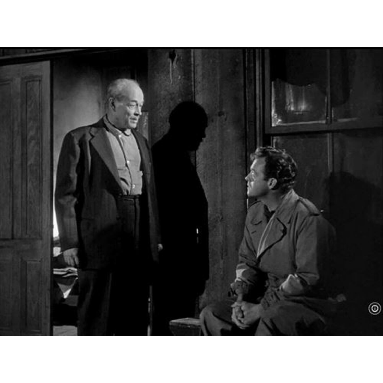 Scène de film en noir et blanc. Deux hommes parlent, un debout, un assis, un autre en arrière-plan.