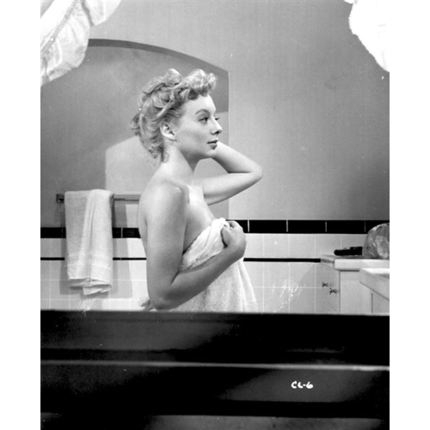Photo en noir et blanc d'une femme dans une salle de bain, drapée dans une serviette, se regardant dans un miroir.