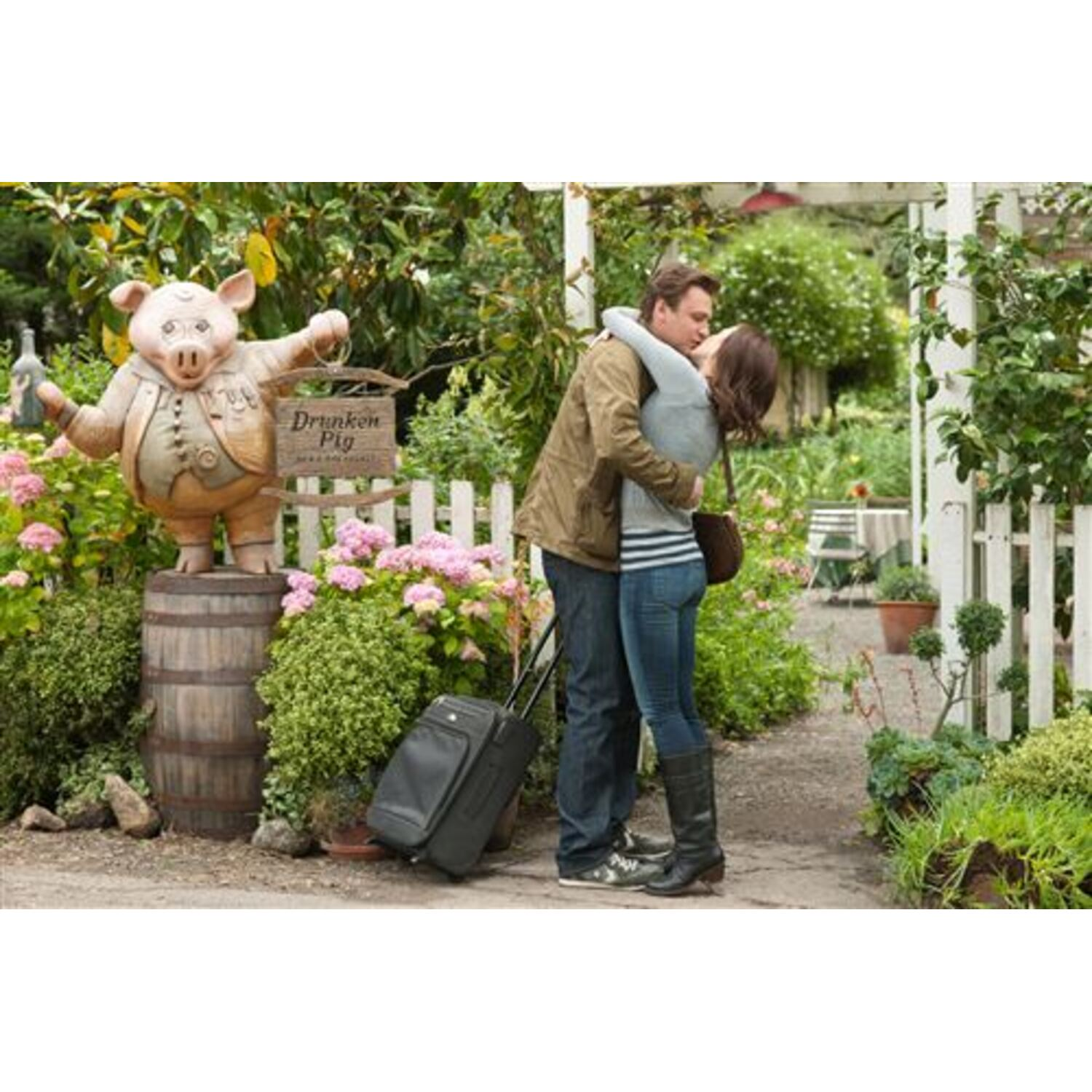Couple s'embrassant dans un jardin, à côté d'une statue de cochon et d'une valise.