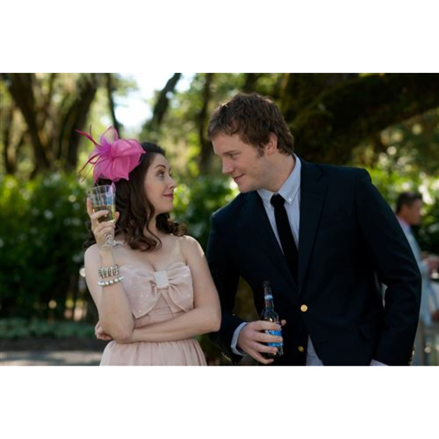 Een man in pak met stropdas met een vrouw in een roze jurk, drankjes vasthoudend, in een park.
