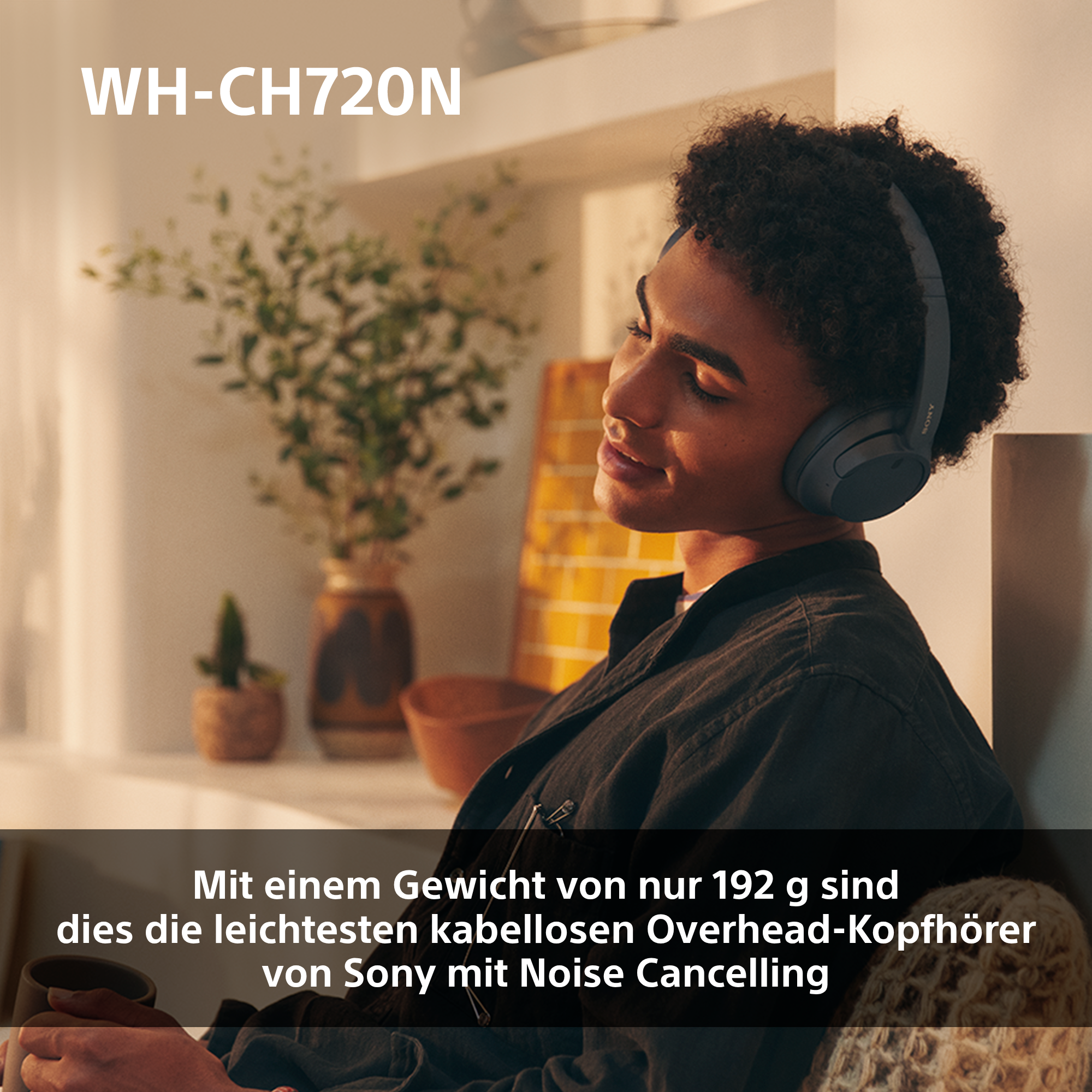 Frau mit grauen Sony Kopfhörern mit Geräuschunterdrückung. Sie lächelt, eine grüne Pflanze ist sichtbar.