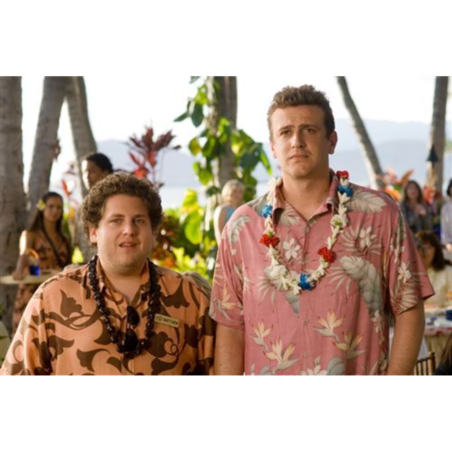 Twee mannen met kransen poseren buiten. Achtergrond met bomen en mensen. Een man draagt een roze shirt.