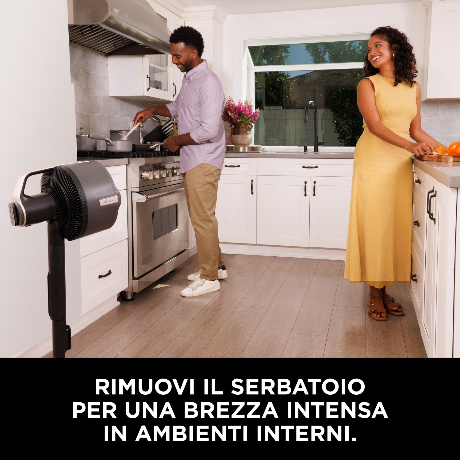 Cucina con una persona che cucina, un'altra in piedi. Ventilatore davanti. Armadi e finestra bianchi sullo sfondo.