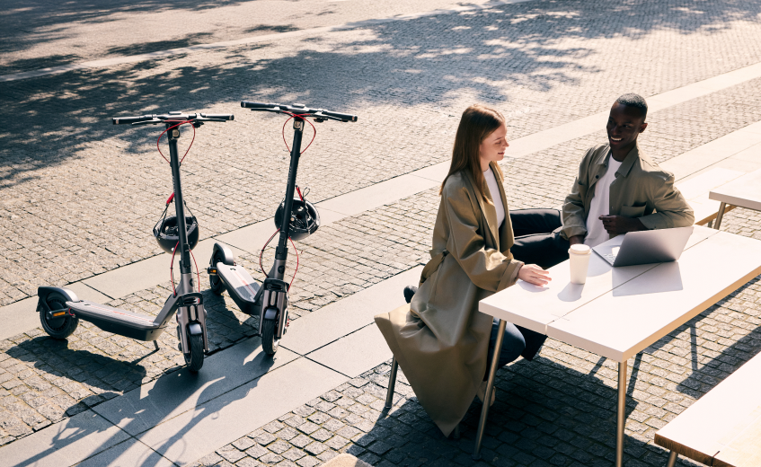 Deux trottinettes électriques et deux personnes à une table avec un ordinateur portable, ensoleillé.